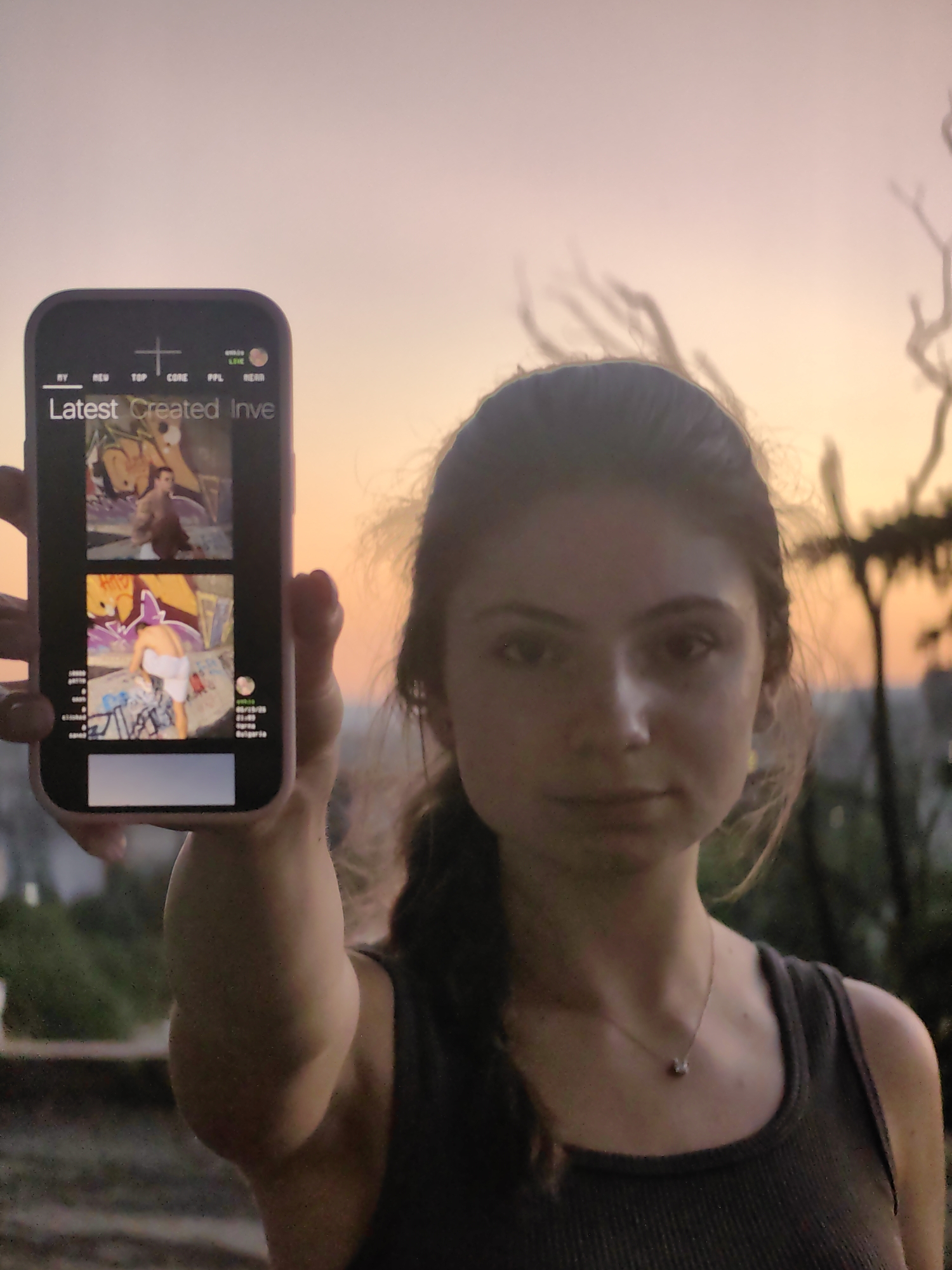 A young woman holds a smartphone displaying a social media feed at sunset in Varna, Bulgaria. The phone screen shows a grid of images, with "Latest Created Inve" at the top. The woman's face is in profile, and she is wearing a dark tank top and a delicate necklace. The background is a soft gradient of orange and pink, characteristic of a sunset, with silhouetted branches of trees visible. The woman's arm is extended, holding the phone forward. The overall scene suggests a casual, outdoor setting in Varna.
