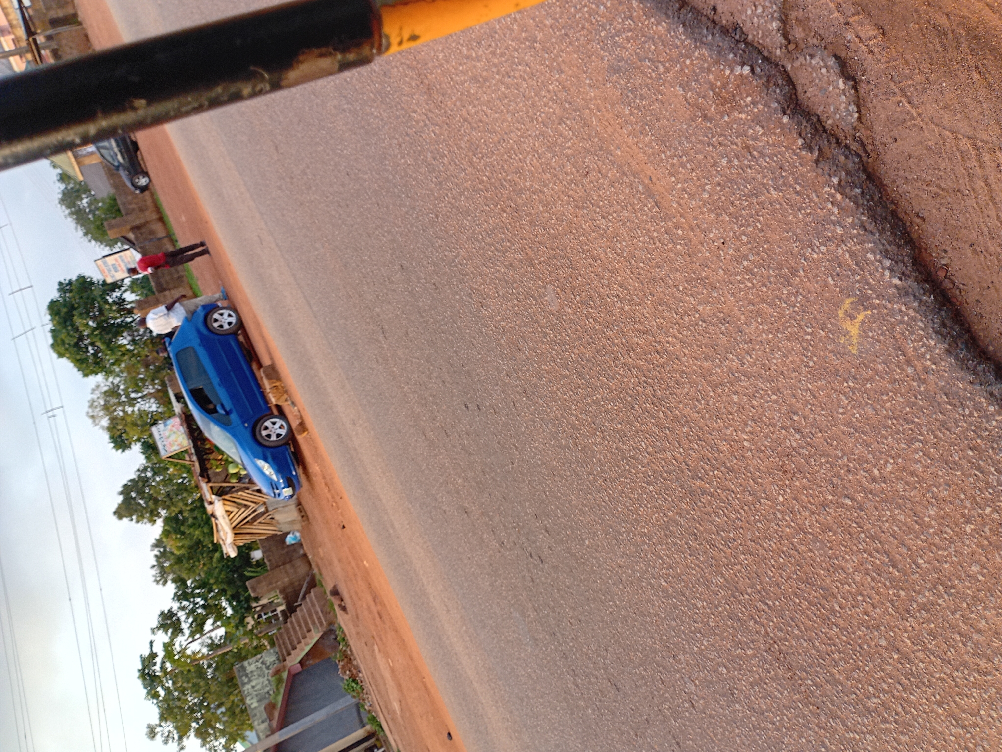 The image depicts a roadside scene in Kuru Dungus, Nigeria, with a clear view of a blue car parked on the side of a dusty, reddish-brown road. Two men are visible near the car. One man, wearing a white shirt and dark pants, stands beside the blue car, while another man in a red shirt and dark pants stands further down the road, facing away from the camera. In the background, there is a small structure with a thatched roof, possibly a vendor stall, and dense green foliage. Another dark-colored car is parked on the opposite side of the road, further in the background. The sky is overcast. The foreground of the image is dominated by the textured surface of the road.