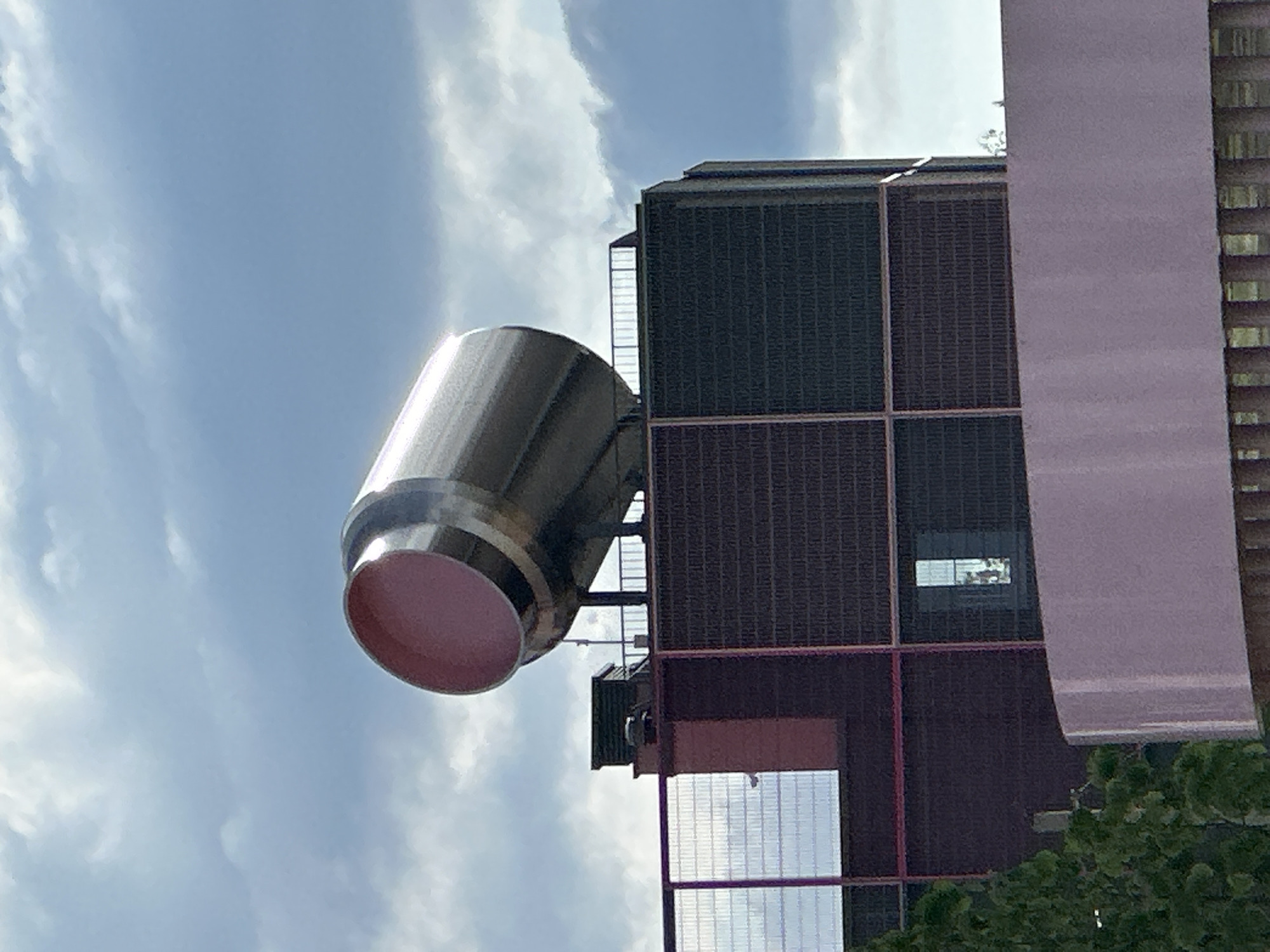This image captures a large, prominent industrial-style cylindrical structure mounted atop a building in Бараўлянскі сельскі Савет, Belarus. The main subject is a substantial, horizontally oriented cylinder crafted from a reflective, brushed metallic material, likely stainless steel or aluminum. Its open end, positioned towards the upper-left, reveals a smooth, matte pink interior. The cylinder is supported by a dark, open metal framework that includes railings, situated on the rooftop of a multi-story building.
The building itself features dark gray or brown facades with a textured surface, possibly composed of brickwork or panels. These dark sections are segmented by reddish-brown vertical and horizontal beams or decorative elements. On the rooftop, directly beneath the large cylinder, a smaller, dark green rectangular unit is visible, alongside metal railings. To the left side of the building, a fine mesh fence partially obscures a white, oval-shaped object mounted on a pole, possibly a surveillance camera or sensor.
In the lower foreground, a smooth, curved structure in a light pink or lavender tone occupies a significant portion of the frame. Below this, a dark railing or fence is partially visible. To the far left, green foliage from a tree or bush is present. The background consists of a bright blue sky interspersed with scattered white and light gray clouds, indicating daylight conditions.
