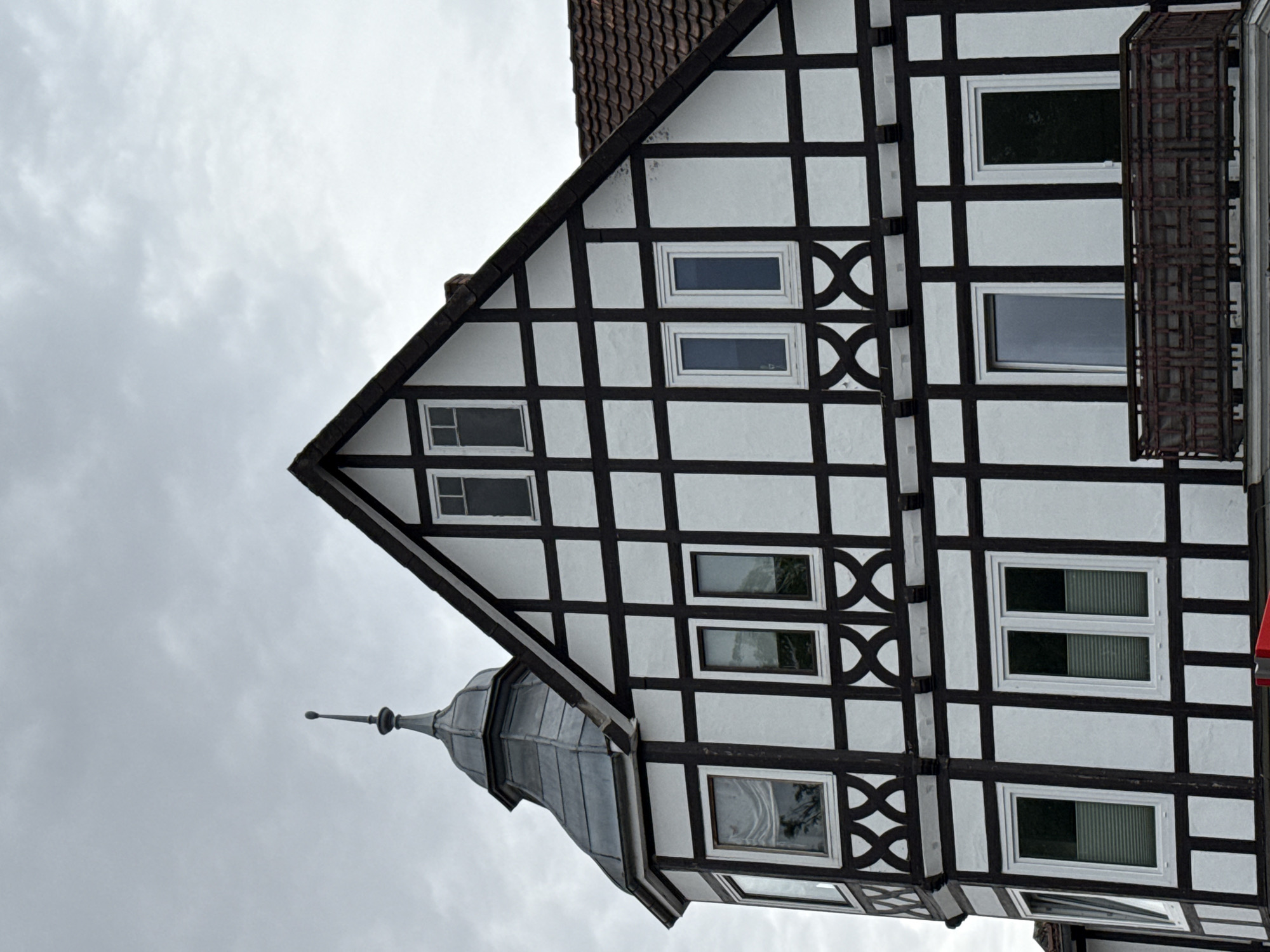 This image shows a close-up of the facade of a historic timber-framed building in Bad Sachsa, Germany. The building features a white stucco exterior with dark brown timber beams forming a geometric pattern. There are multiple windows of various sizes, some with white frames and others with darker panes. A distinctive turret with a conical, dark metal roof and a pointed finial rises from the left side of the building. The roofline of the main structure is tiled in a reddish-brown color. A wrought-iron balcony is visible on the lower level, projecting from the building. The sky above is overcast with diffused light.