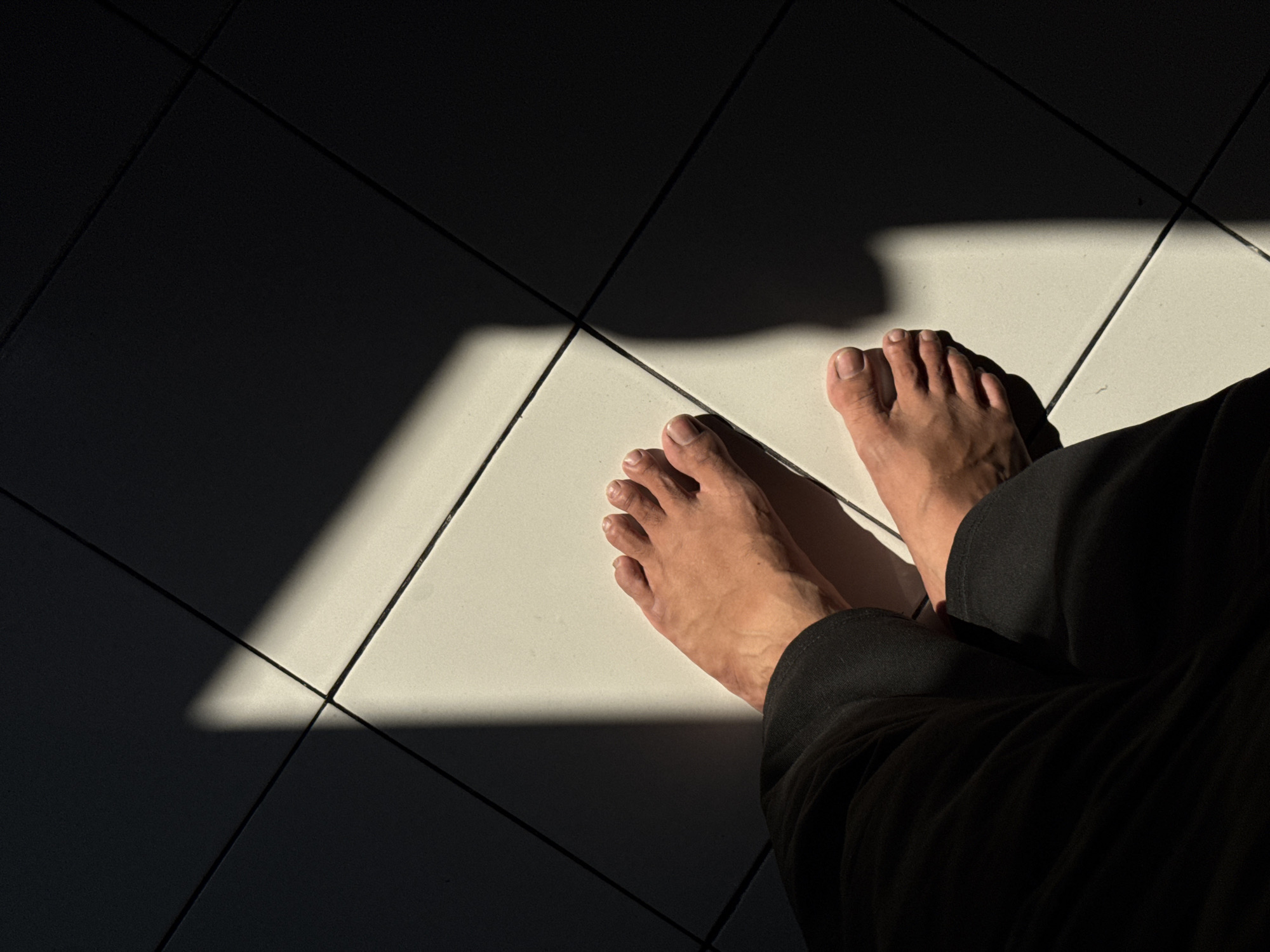 The image shows two bare feet positioned on a tiled floor, with a sharp diagonal shadow cast across them. The tiles appear to be light-colored and square, arranged in a grid pattern. One foot is closer to the viewer, with toes clearly visible, and is partially covered by dark fabric, likely pants. The other foot is further away and mostly illuminated by sunlight. The lighting suggests a strong light source, possibly from a window or an opening, creating a high contrast between the illuminated and shadowed areas of the floor. There are no other people or objects visible in the frame. The scene is likely indoors or in a covered outdoor space due to the tiled floor and the nature of the shadow. The location is specified as Kalirancang, Indonesia.