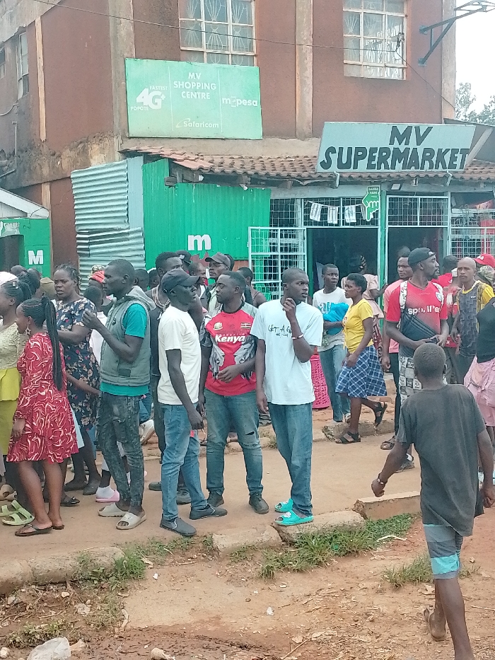 The image shows a crowd of people outside of a supermarket in Kivaywa, Kenya (latitude: 0.562, longitude: 34.805). The supermarket is named "MV Supermarket" and has a sign above the entrance that reads "MV Shopping Centre m-pesa Safaricom." There is a green corrugated metal awning covering the storefront. There are at least 20 people in the image, most of whom are standing in a line outside the supermarket. There are multiple people standing inside the entrance of the supermarket. Some are wearing hats and backpacks. The people in the crowd are interacting with one another, seemingly waiting to enter the store. The ground is dirt and there is some greenery in the foreground of the image. The store appears to be in an urban location with houses visible in the background.
