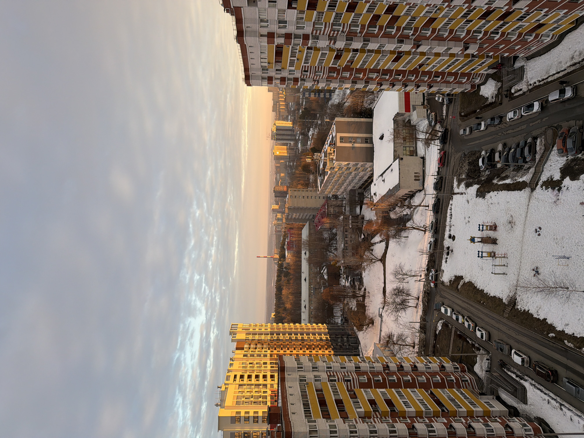 An aerial view captures a winter or early spring urban landscape in Izhevsk, Russia, under cloudy skies with low-angle golden hour sunlight. The scene is dominated by residential high-rise buildings and snow-covered terrain.
In the immediate foreground, a multi-story residential building with white, yellow, and red-brown facade panels occupies the lower left. To its right, partially obscured, is a snow-covered road with approximately ten parked cars. A children's playground, featuring colorful slides and climbing structures, is situated on a snow-covered patch of ground in the lower center. To the lower right, another multi-story residential building, mirroring the design of the one on the left, stands prominently, partially shadowed. A winding road with approximately fifteen parked vehicles runs along the base of this building. At least two indistinct human figures are visible on the sidewalk adjacent to the building and on the road.
The midground features an expanse of partially snow-covered ground with numerous leafless deciduous trees. Scattered throughout are various multi-story buildings of differing architectural styles and heights, some with flat roofs and visible communication equipment. Roads traverse this area, with more parked cars.
The background presents a vast cityscape extending to the horizon. Buildings appear progressively smaller and more densely packed. A prominent red and white striped industrial chimney rises above the skyline to the left of center. A significant forested area is visible towards the right midground, providing a natural buffer within the urban environment. The distant sky and cityscape are bathed in a warm, golden-orange light from the low sun, creating a hazy horizon line beneath a sky with stratified grey and white clouds.
