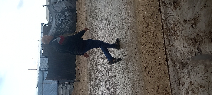 The image depicts an outdoor scene in Khan Yunis, Palestinian Territory, featuring a single adult male walking from left to right across a wet, muddy, unpaved ground surface under an overcast sky. The man, balding with short light-colored hair, wears a black long-sleeved jacket with a red and white diagonal stripe pattern on the left shoulder, dark blue trousers, and dark open-toed sandals. He is in mid-stride, with his left foot lifted off the ground and his arms in natural walking motion.
In the background, a row of makeshift shelters or structures is visible. These structures are constructed from various materials including dark and grey tarpaulins and blankets, stretched over visible frames. One prominent dark blanket on a shelter in the right background displays a distinctive white or light-colored pattern resembling floral or abstract designs. The ground directly behind the man appears to be packed earth. The foreground shows a darker, more saturated section of wet ground or possibly a different material, delineated by a horizontal line from the muddier, reflective middle ground.