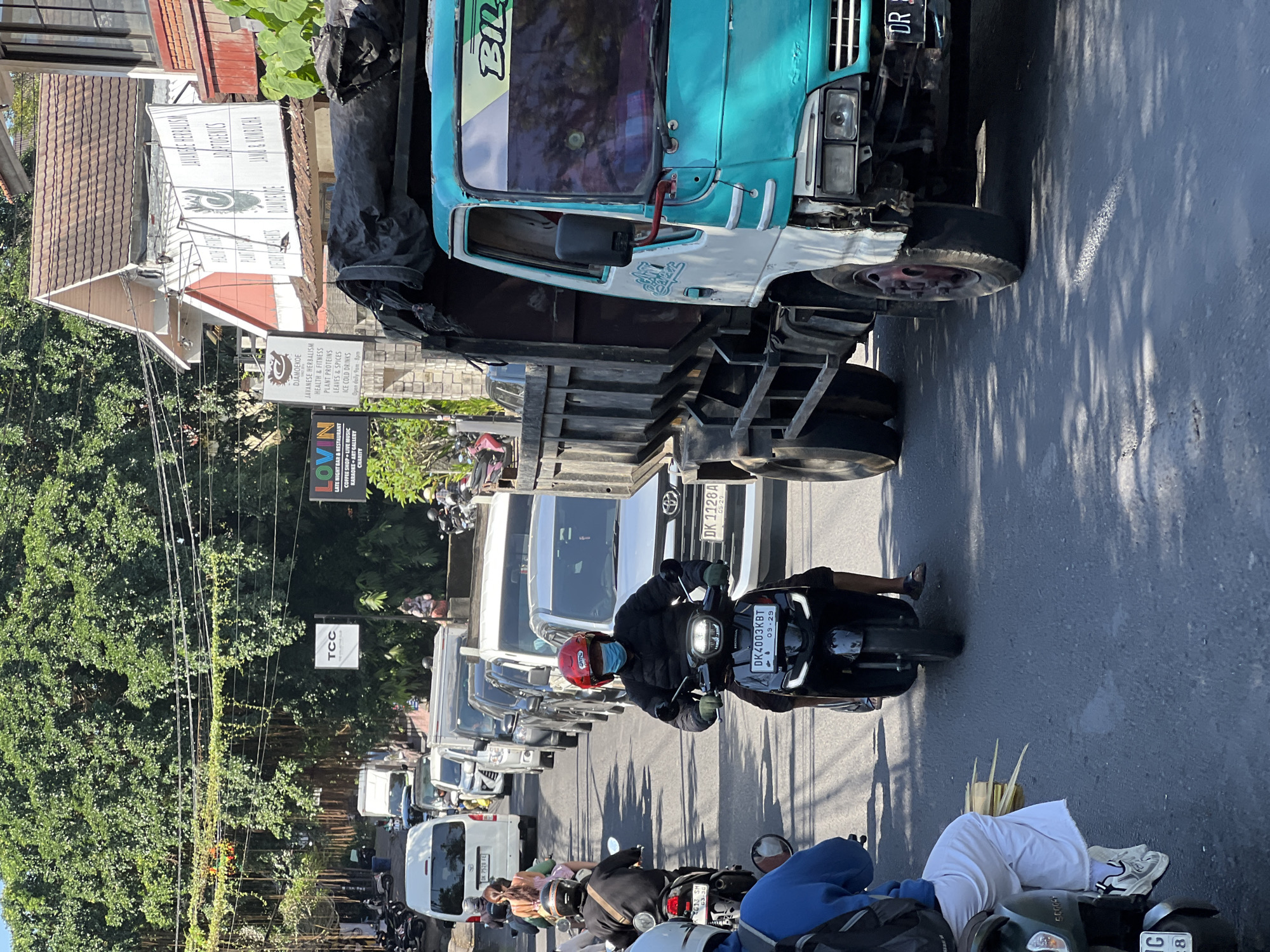 The image depicts a traffic jam on a street in Ubud District, Indonesia, on a sunny day. A turquoise and white dump truck with a black tarp covering its load occupies the right side of the frame. In front of the truck, several motorcycles and cars are stopped. A man wearing a red helmet and a blue face mask rides a black scooter in the foreground, with a license plate reading "DK 4003 KBT". He is wearing a black jacket and green gloves. Behind him, another person on a motorcycle wears a blue jacket and white pants, with palm fronds protruding from the side of their vehicle. To the left of the truck, a white van is visible. Buildings and lush green trees line the street, with hanging vines and power lines overhead. Signage for businesses like "TCC" and "DJAMOEKoe" are present on the buildings.
