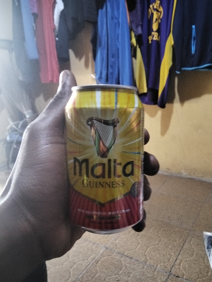 The image shows a dark-skinned human left hand holding a yellow and red can of Malta Guinness, a non-alcoholic malt drink, in Badore, Nigeria. The can features a prominent harp logo in green, orange, and white, with "Malta GUINNESS" text displayed centrally. The bottom section of the can explicitly states "NON ALCOHOLIC MALT DRINK," and a silver pull tab is visible at the top. The background depicts an indoor environment with a solid yellowish-cream colored wall. Several items of clothing, including blue shirts, a pink shirt, dark garments, and a purple garment with yellow accents, hang from a rack or line behind the can. A light-colored tiled floor with a repeating embossed pattern is partially visible in the bottom right foreground.