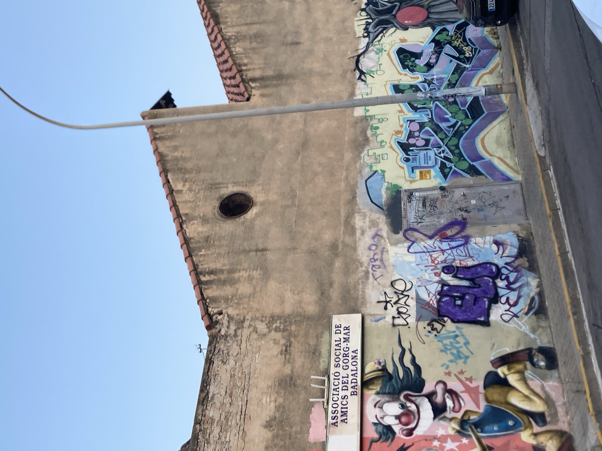 The image shows a section of a building facade adorned with vibrant graffiti. On the left side of the facade, a sign reads "ASSOCIACIÓ SOCIAL DE AMICS DEL GORG-MAR BADALONA," indicating this is the Gorg-Mar Social Association in Badalona, Spain. Below the sign, a colorful mural features a cartoonish clown character in motion, with exaggerated features and attire.
The wall is extensively covered in graffiti, displaying various styles and colors. Prominent graffiti pieces include stylized lettering in purple and blue, along with abstract shapes and patterns in green and purple. A tall, slender streetlight stands in front of the wall, partially obscuring some of the graffiti. To the right, a dark-colored tree-like figure with a large red element, possibly an eye or fruit, is depicted in graffiti. A portion of a black car with license plate "224" is visible on the far right.
The building itself appears aged, with weathered walls and a tiled roof. A circular opening, resembling a small window or vent, is visible high on the wall. The foreground shows a sidewalk and a street with yellow and white markings. The sky is clear and blue. The overall scene depicts an urban environment characterized by street art and the architectural features of an older building.