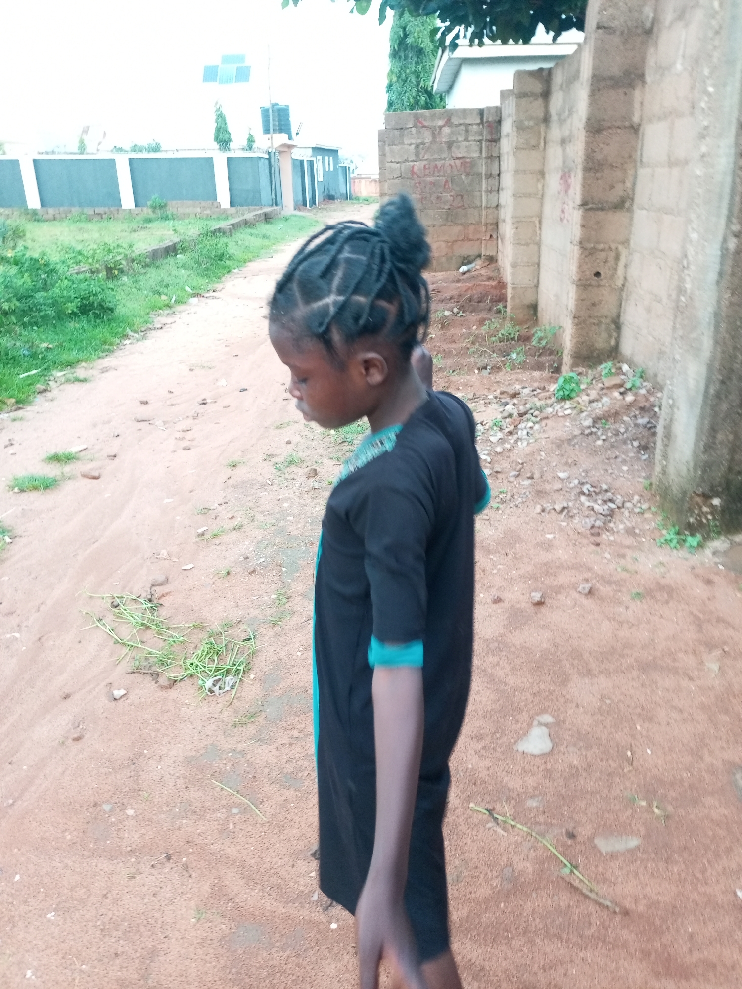 In Tempure, Nigeria, a young Black girl with braided hair pulled into a bun stands on a dusty dirt path. She is wearing a black dress with turquoise trim. The path is lined with sparse green vegetation and scattered stones. In the background, a brick wall with graffiti is visible, along with a building and what appear to be solar panels. The environment suggests an outdoor, possibly semi-rural or developing urban setting.