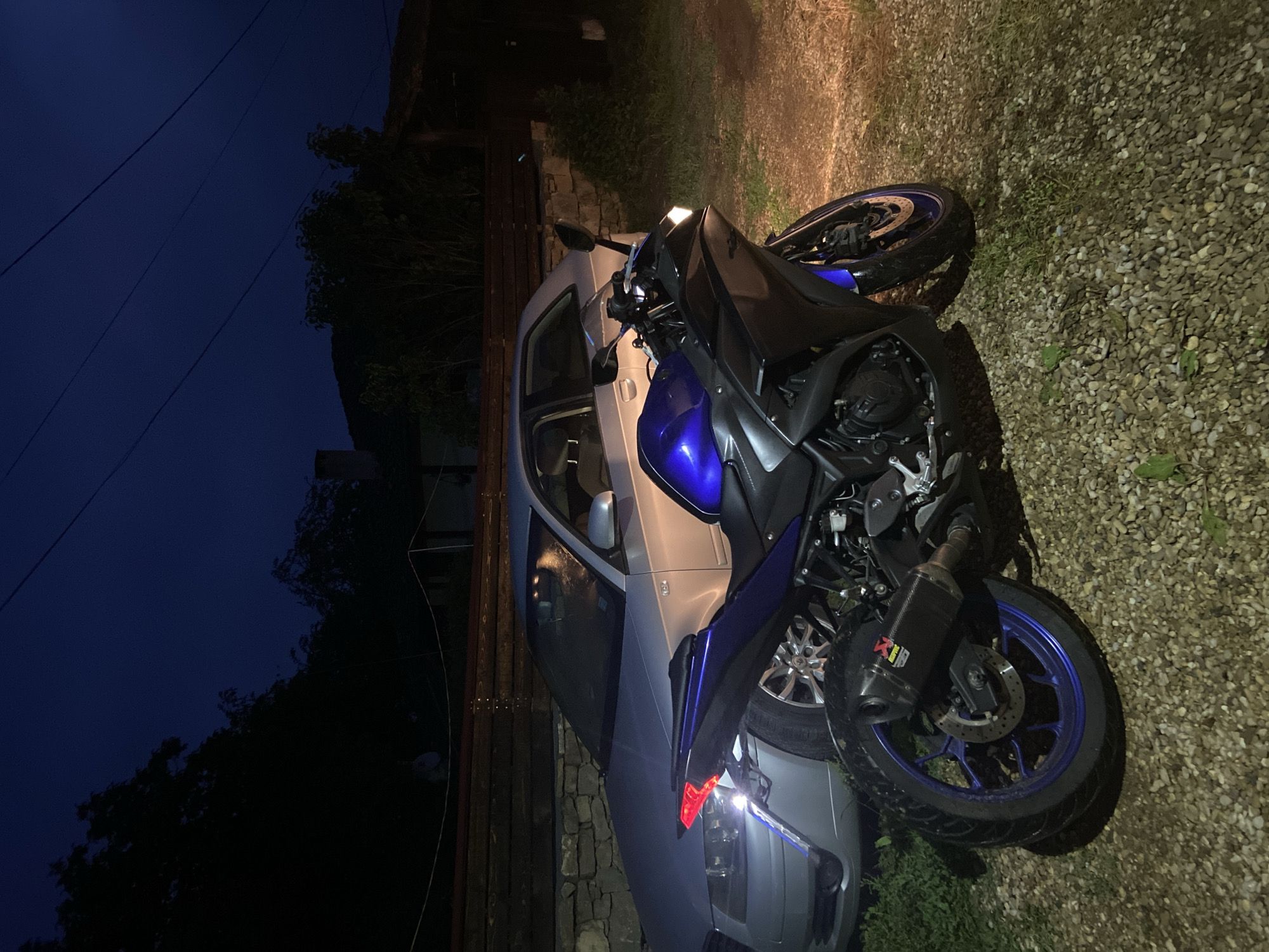 The image, captured at night, presents a static scene featuring a blue and black motorcycle parked in front of a silver or light-colored car. Both vehicles are situated on a gravel surface. The car's headlights are illuminated, casting a bright light on the foreground, revealing details of the gravel and the front section of the motorcycle. The car's left rear tail light also glows red.
The motorcycle, a sport bike, features prominent blue accents on its fairings and wheel rims, contrasting with its black bodywork. An aftermarket exhaust system is visible, clearly displaying the brand name "Akrapovič" and its logo on the carbon fiber-textured canister. The front wheel of the motorcycle is turned towards the car.
In the background, a dark blue night sky dominates the left side, traversed by thin overhead wires. On the right, silhouetted trees and dense foliage are visible against the dark sky, suggesting a natural or semi-rural setting. A wooden fence and a stone wall run alongside the gravel area, further defining the location. No people are visible in the image. The scene conveys a sense of quiet stillness at night, likely in a private parking area or driveway in a location such as Mindia, Bulgaria.