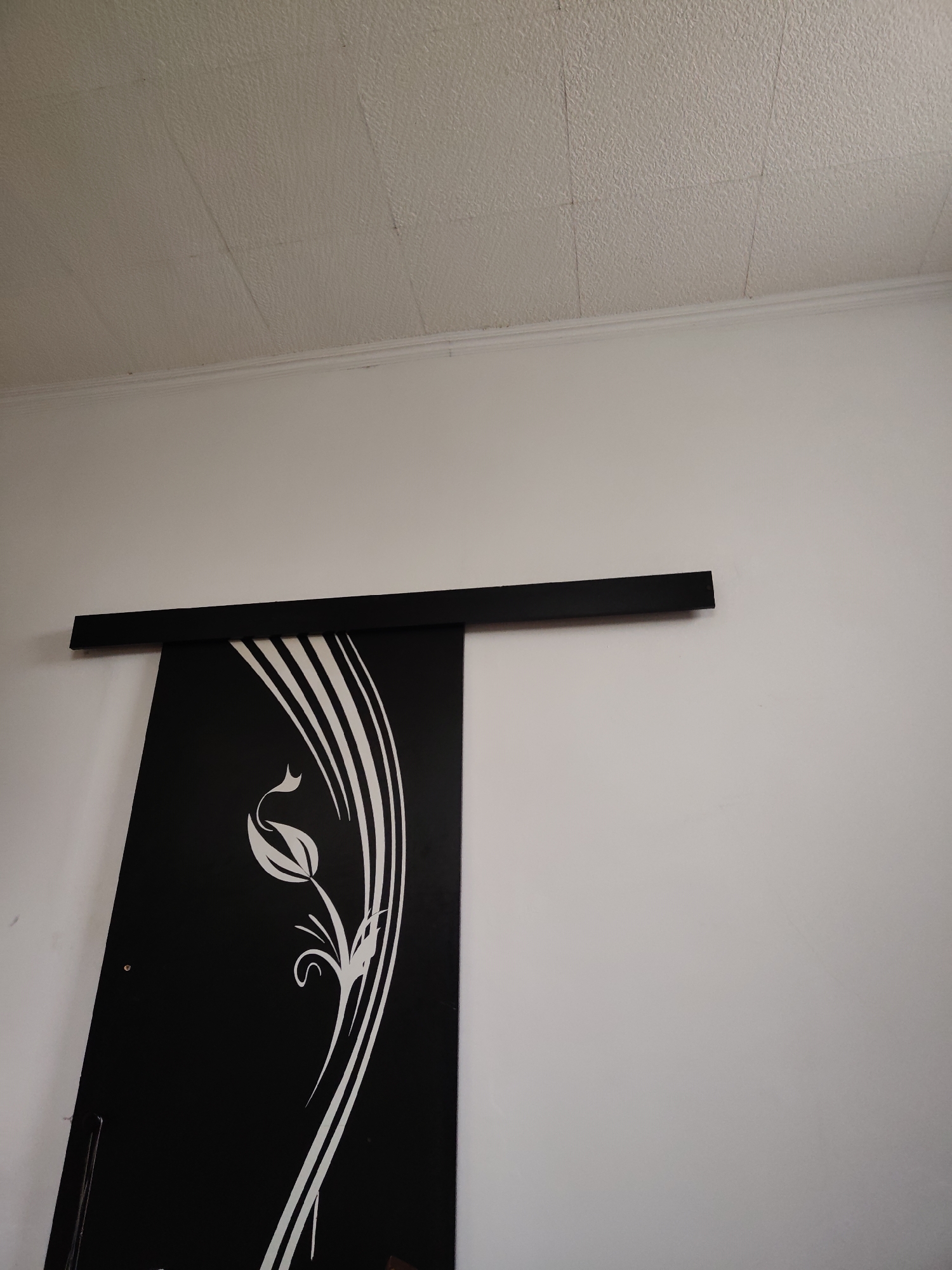 The image shows a black sliding door with a white floral design, mounted on a wall. A black rail is visible above the door. The ceiling is textured and white, with a white crown molding along the top of the wall. The image appears to be an interior shot of a room. No specific location in Dobiegniew, Poland is identifiable from the image.