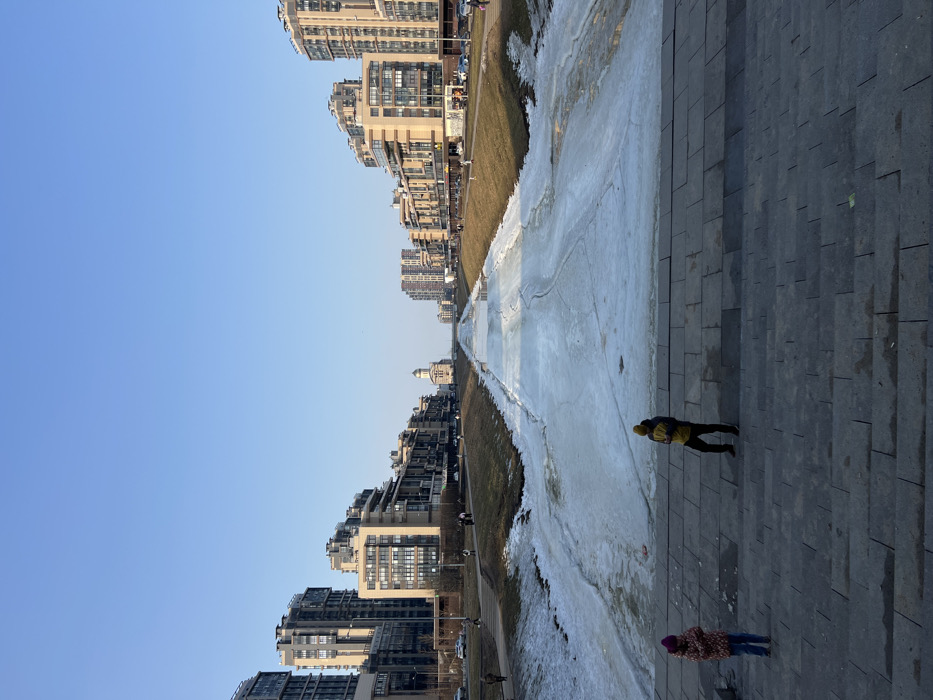 The image depicts an outdoor urban scene featuring the frozen Smolenka River on Vasilyevsky Island in Saint Petersburg, Russia, with the Church of St. Michael the Archangel visible in the distance. The view is from a multi-tiered paved staircase descending towards the river.
The river, running centrally through the image, is largely covered by white, fractured ice, indicating a thawing state, with some patches of dark water visible. The banks on either side are sloped and covered with sparse, brown-green grass and residual snow.
On the left side of the foreground, a young individual, possibly a child, stands on the stone steps, wearing a pink or purple beanie and a multi-colored jacket. To their right, an adult figure stands on a lower step, wearing a yellow beanie, a yellow and dark jacket, and dark pants. Both individuals face forward, towards the river.
Lining both sides of the river are multi-story modern residential buildings, typically 10 to 15 stories tall, featuring light-colored and dark facades with numerous windows. These buildings extend into the midground and background, creating an urban corridor along the waterway. Roads or pathways run parallel to the riverbanks, with several smaller figures of people walking and a few vehicles visible.
In the far distance, centered along the river's axis, stands the Church of St. Michael the Archangel, characterized by its light-colored dome and traditional architectural elements. The sky above is clear and pale blue. The overall impression is of a late winter or early spring day.
