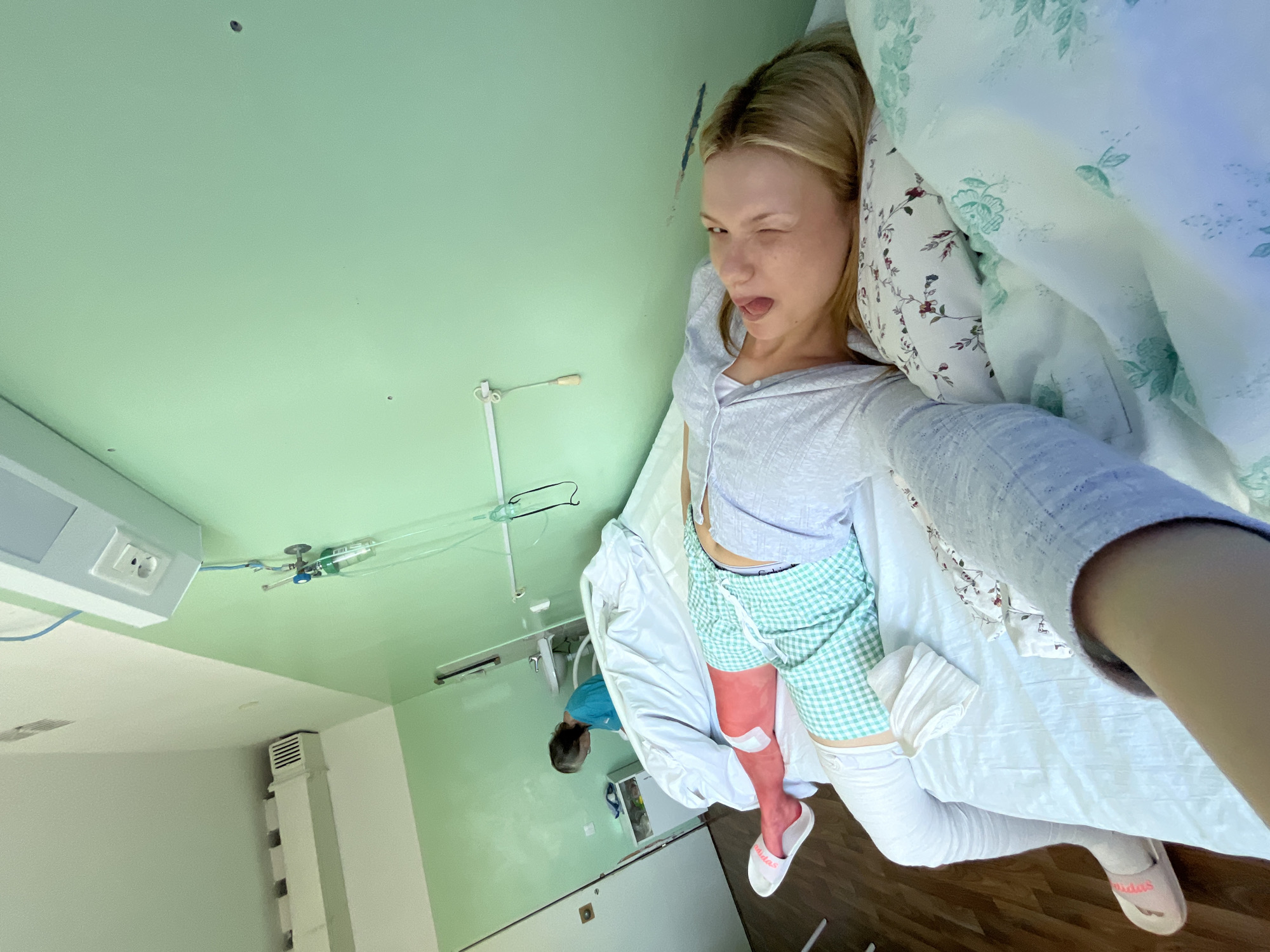 A woman is reclining in a hospital bed in a room with green walls and a white ceiling in Chaikovsky, Russia. She is fair-skinned with blonde hair, and wearing a grey shirt, green checkered shorts, and white compression stockings on both legs. Her right leg is red and bandaged. She is making a silly face, winking and sticking her tongue out, appearing to take a selfie. The bed has white sheets and a floral-patterned comforter. An individual in a blue shirt is bent over in the background near medical equipment. The room is sparsely furnished and contains standard hospital equipment, including medical gas outlets and an examination light on the wall.