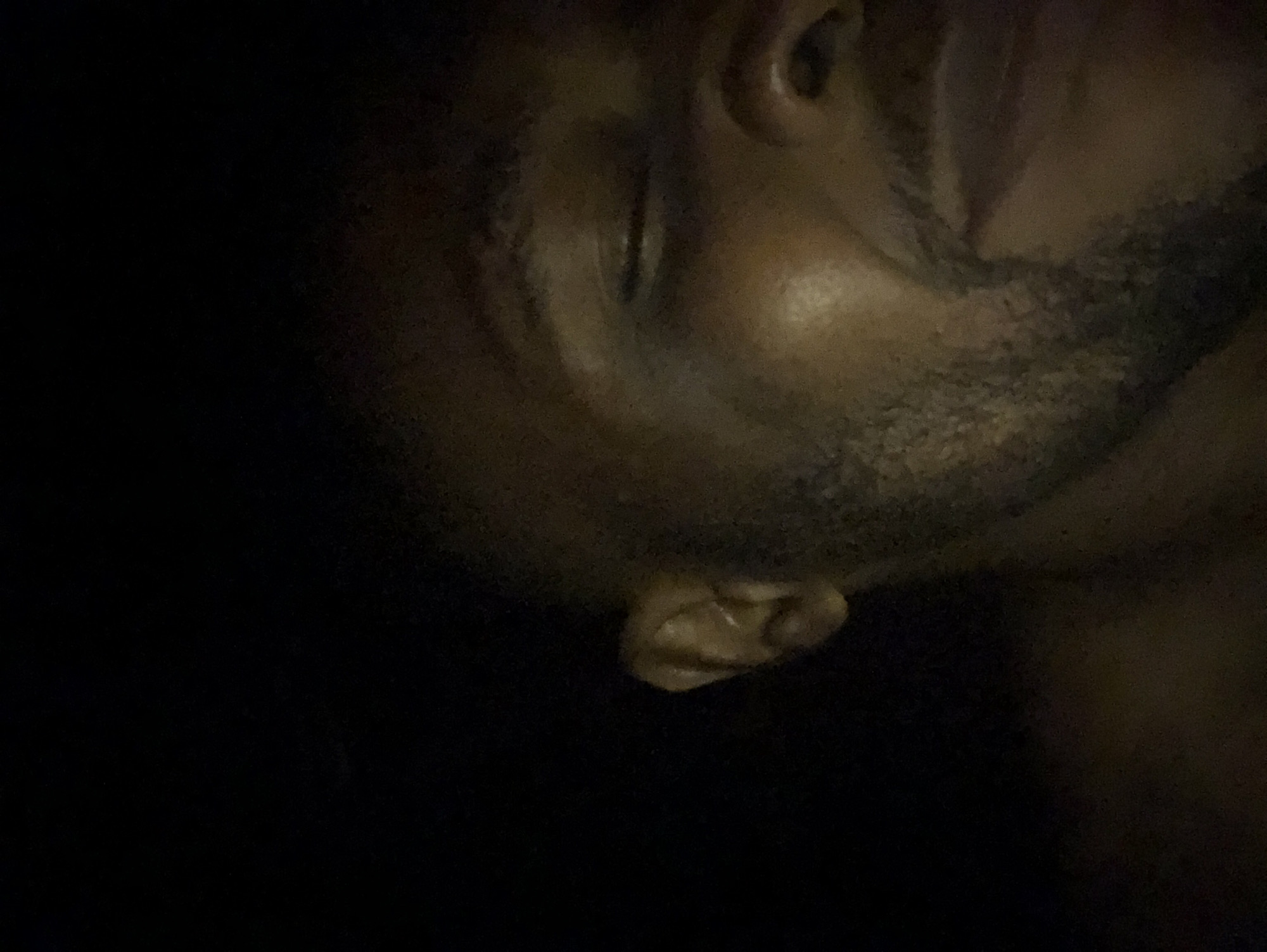 The image is a close-up of a man's face in low lighting. The man has dark skin, a closed eye, and a beard. Part of his ear is also visible. The background is dark and indistinct. The location is unknown.