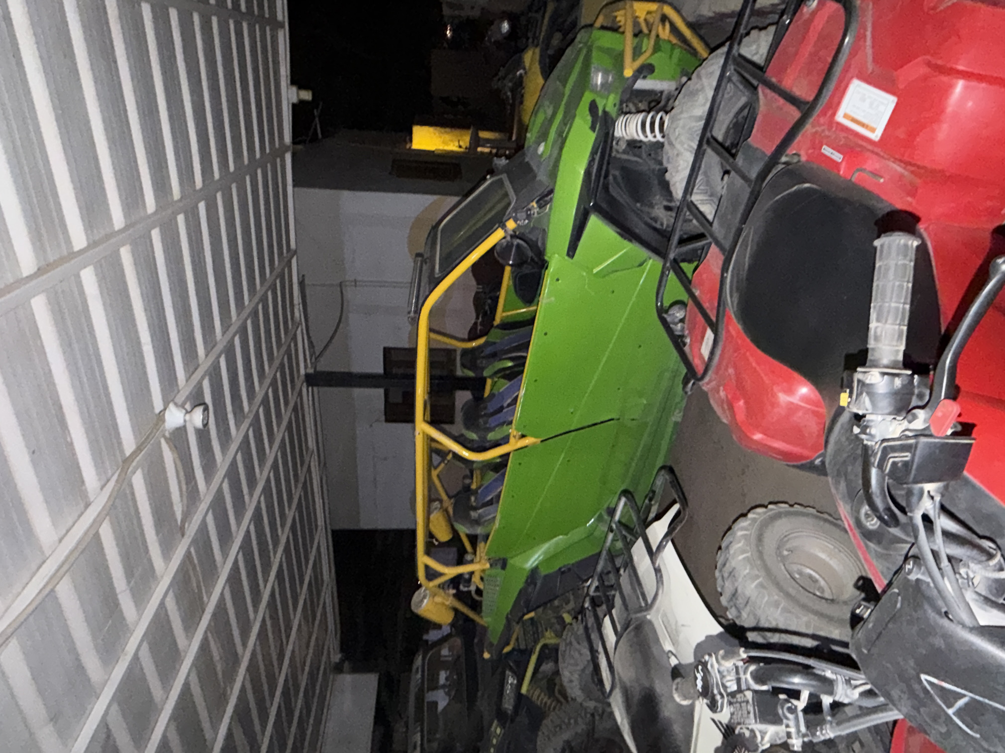 This image depicts an indoor scene, possibly a garage or storage area, under a corrugated metal roof supported by a metal frame. The lighting is artificial, casting shadows. In the foreground, two red ATVs are parked. The ATV on the right is partially visible, showing its rear section with a black seat and a metal rack. The ATV on the left is more prominently displayed, with its front wheel and handlebars visible, along with parts of its body.
Dominating the middle ground is a bright green off-road buggy with a yellow roll cage. The buggy has a two-tone interior with dark seats. It appears to be parked with its front end facing towards the right of the frame, and its side is clearly visible. Another dark-colored off-road vehicle is partially obscured behind the green buggy.
The overall environment is cluttered with these recreational vehicles. The image does not provide any direct visual cues to pinpoint its exact location within Sharm El Sheikh, Egypt. There are no people visible in the scene, and no specific actions or interactions are taking place. The scene is static and appears to be a storage or staging area for these vehicles.