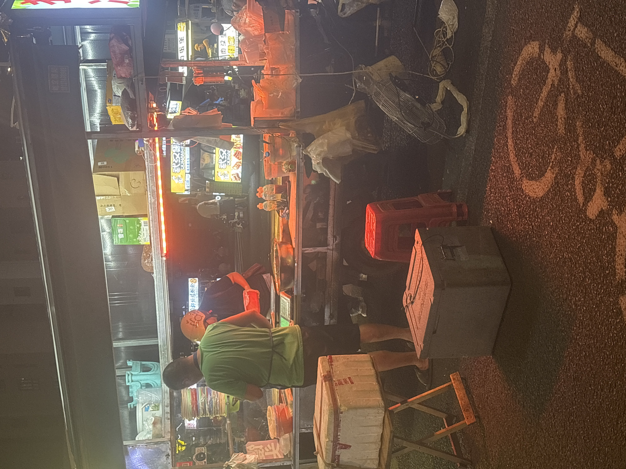 The image captures a nighttime scene at a food stall in Suzhou Industrial Park, China. The food stall is illuminated by red and yellow lights, with various food items and cooking equipment visible. Two individuals are present: one man in a green shirt and black shorts is standing behind the counter, appearing to be preparing food or serving customers, and another individual wearing a helmet is also behind the counter. Boxes and containers are stacked on shelves and on the ground, including what appears to be a large styrofoam cooler in the foreground. A red stool and a bicycle symbol painted on the ground are also visible. The overall environment suggests a busy urban setting at night.