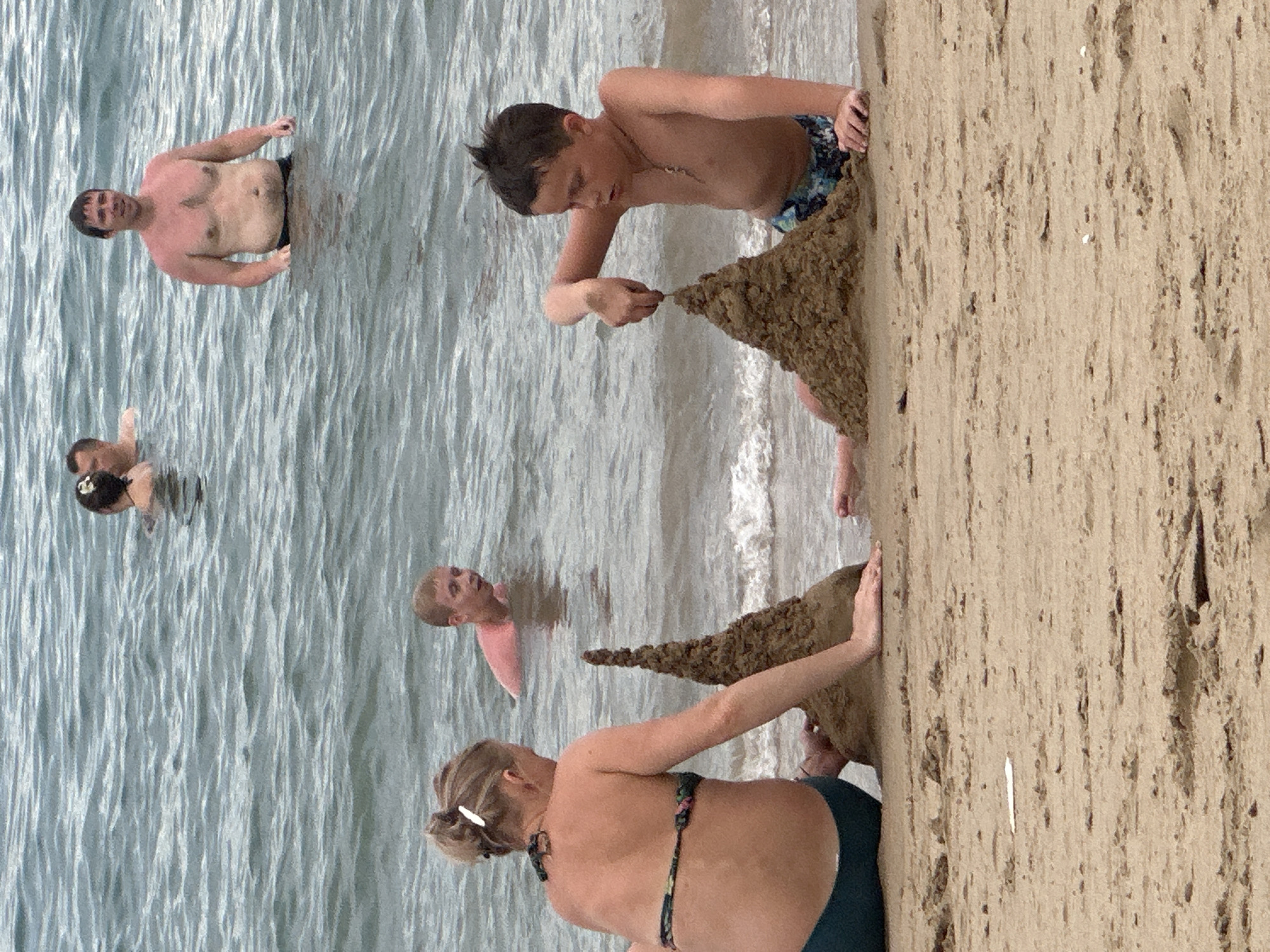 The image depicts a vibrant beach scene in Nha Trang Ward, Vietnam, showcasing individuals enjoying leisure activities during what appears to be a bright daytime.
In the foreground, a young boy with dark hair is intensely focused on building a large, conical sand structure near the water's edge. He is shirtless, wearing patterned swim shorts, and is actively shaping the sand with his hands. Behind him, a woman with blonde hair, styled in a bun and wearing a two-piece swimsuit, is also interacting with the sand structure, kneeling on the beach.
Further out in the calm ocean water, several other individuals are visible. A man with short dark hair, wearing dark swim trunks, stands waist-deep in the water, looking towards the shore. Below him, two other people are partially submerged, including a woman with dark hair and another individual whose head is just above the surface. To the right, another person with light hair and a pink top is seen in the water.
The scene conveys a relaxed and enjoyable atmosphere, typical of a day at the beach, with people engaged in both swimming and sand play. The sand is light brown, and the water appears clear with gentle waves near the shore. No text is visible in the image.