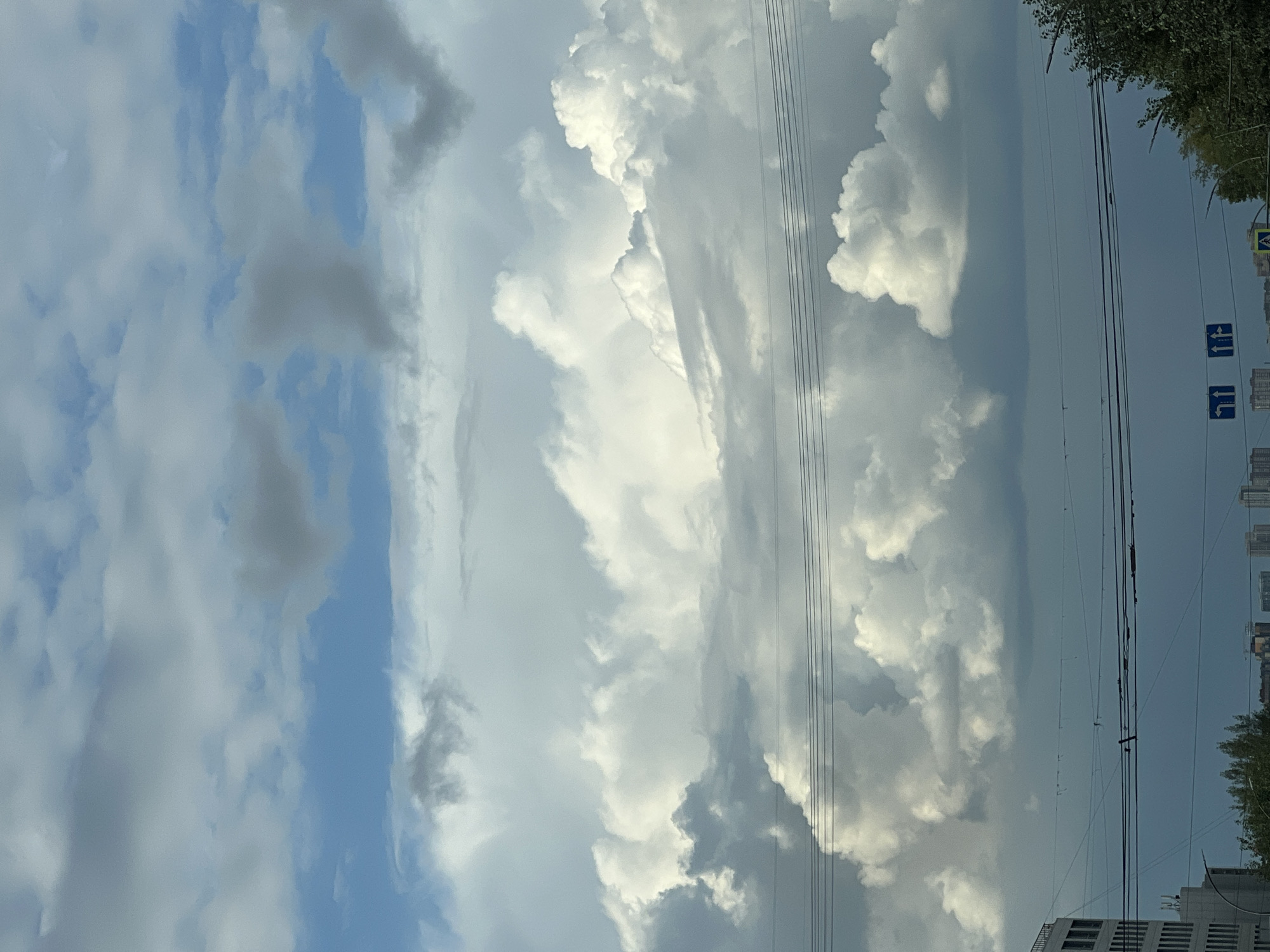 The image shows a cloudy sky with some sunlight filtering through, suggesting overcast conditions over a city. In the foreground, there are multiple power lines stretching horizontally across the frame, with a building on the left side and trees on the right. Below the power lines, street signs with directional arrows are visible, indicating an intersection or road network. The overall scene depicts an urban environment under a dynamic sky. Without specific landmarks or identifiable architectural styles, it is not possible to confirm the location as Novosibirsk, Russia.