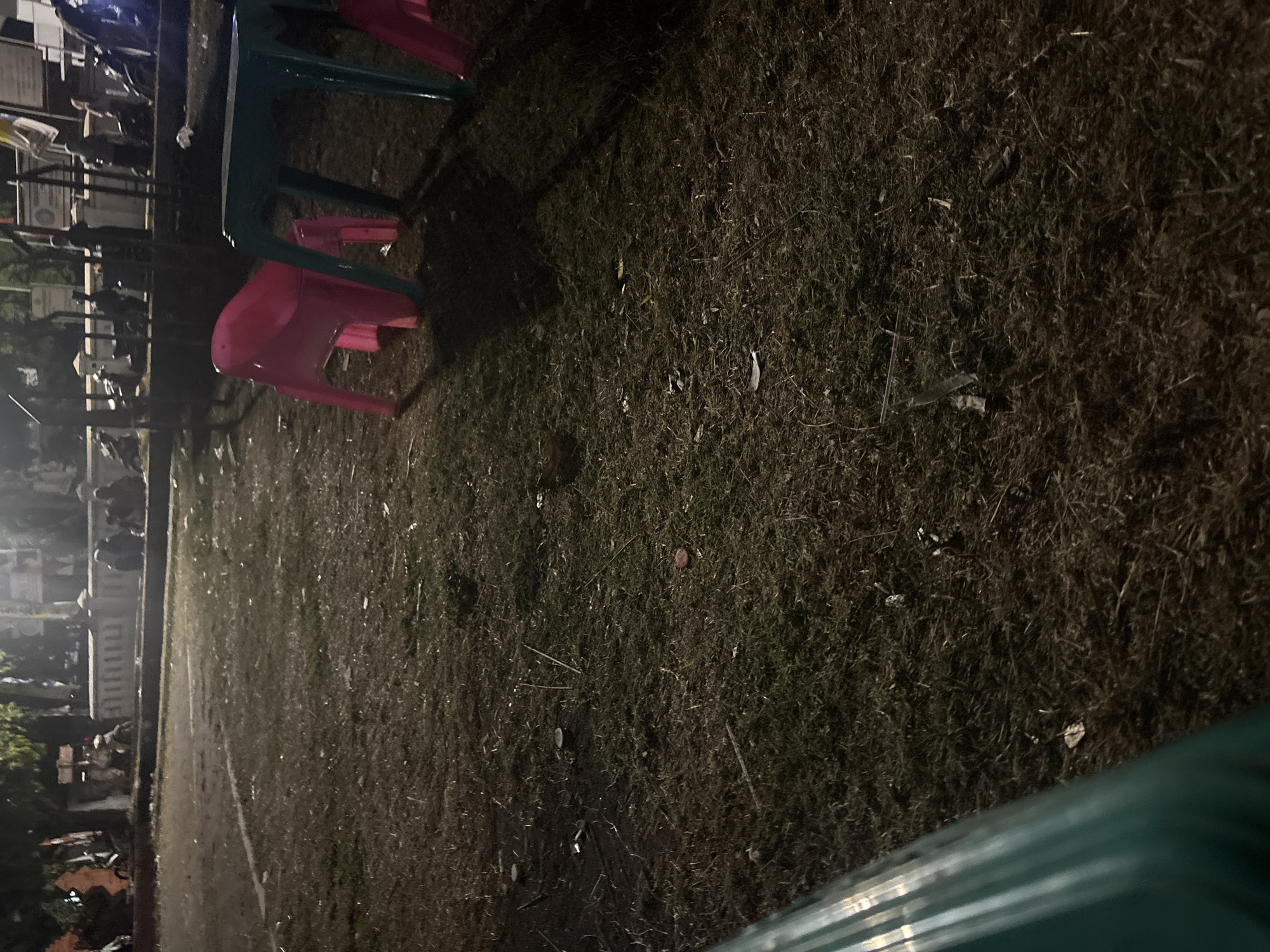 This is a nighttime outdoor scene at a park or open space in Palimanan, Indonesia. The ground is covered in dry, sparse grass and debris. In the foreground, a portion of a blurry green object, possibly a railing or structure, is visible on the left. Further back, two plastic chairs are prominent: one pink chair on the left and a teal table with legs on the right. Behind the chairs, a low, white, decorative wall or barrier is visible, separating the grassy area from a paved pathway or road. A group of people and motorcycles are gathered near this wall, with some individuals seated and others standing. The overall lighting suggests it is dark, with artificial lights illuminating parts of the scene.