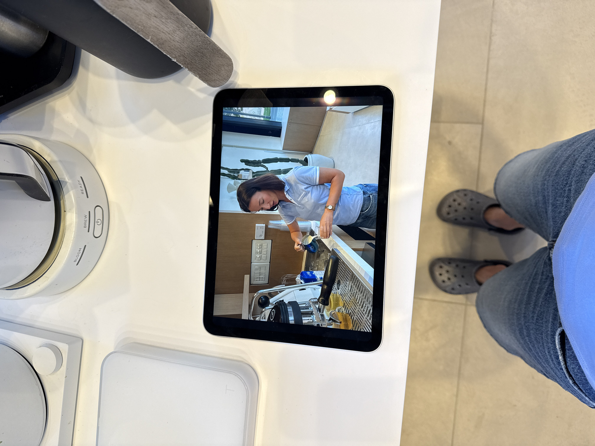 A first-person perspective image depicts a person standing on a light-colored tiled floor, wearing blue jeans and dark gray Crocs. The subject is looking down at a clean, white countertop. On the countertop, a tablet with a black bezel and silver edge displays a video.
The video shows a woman with dark brown hair, wearing a light blue polo shirt, blue jeans, and a watch on her left wrist. She is performing an action consistent with latte art, pouring frothed milk from a silver pitcher into a small white cup, next to a lever-operated espresso machine. Two transparent vessels containing an amber liquid are visible on the machine's drip tray. The background of the video reveals a modern interior with light-colored walls, wooden cabinetry, large windows, and tall potted cacti. A sign partially visible in the background displays "NEXT 9".
On the white countertop, behind and to the sides of the tablet, various white kitchen appliances are present. These include a white coffee maker with "BLOOM," "BREW," and "READY" indicators, and a power button. Another white appliance with a circular knob is visible to the left. A dark gray or black object is partially visible on the right side of the countertop.
Based on the "NEXT 9" signage visible in the video displayed on the tablet, this scene is located at the NEXT 9 coffee bar and showroom, situated at Rothschild Boulevard 9, Tel-Aviv, Israel.