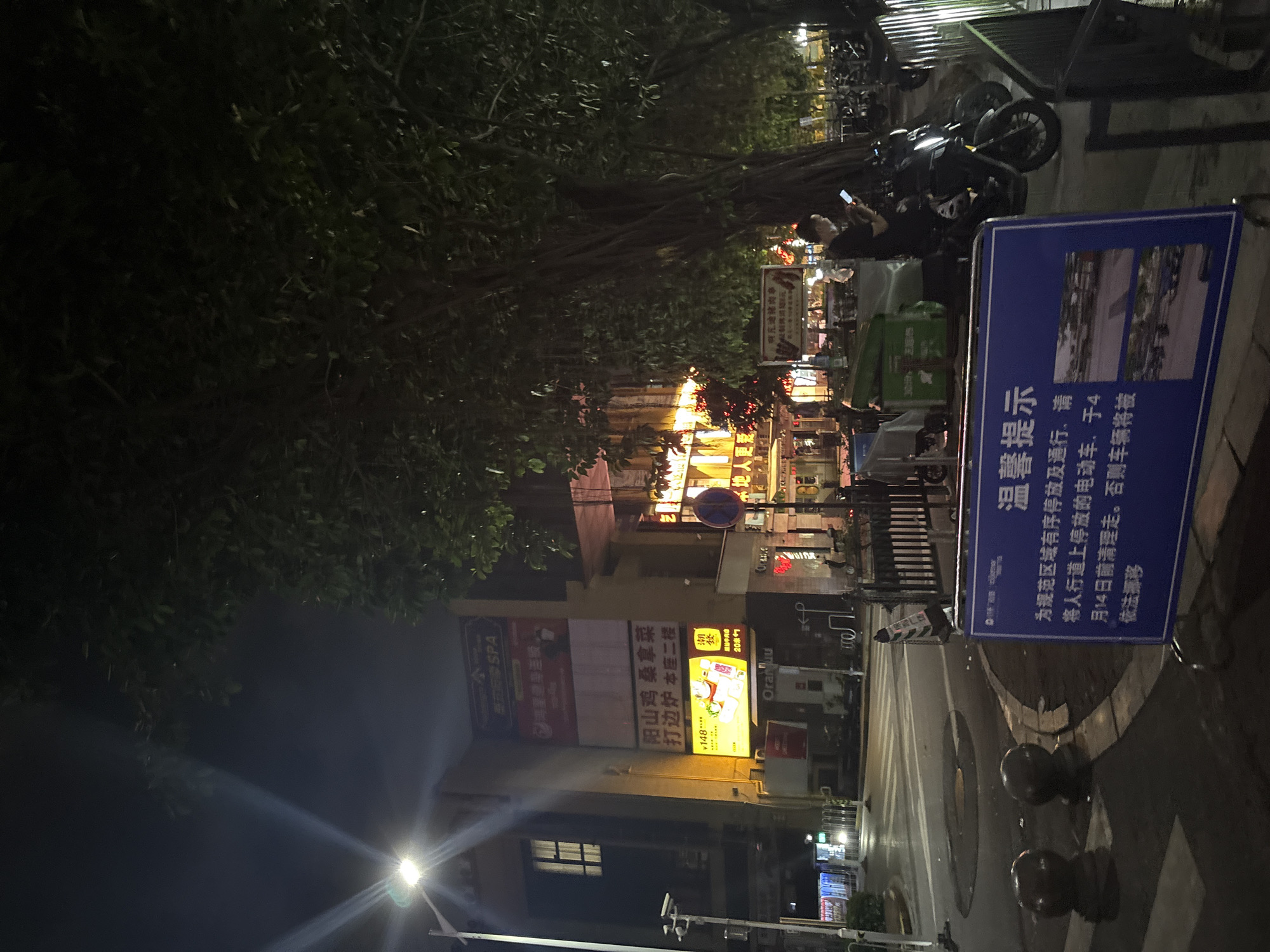 This is a medium-shot, slightly tilted photograph taken at night in Haizhu District, China, depicting a street scene with commercial establishments and a public notice. The primary subjects are the storefronts of various businesses, illuminated by bright, colorful signs, and a blue informational sign with Chinese text.
The setting appears to be an urban street at night, evidenced by the artificial lighting and the presence of parked motorcycles and a delivery cart. The atmosphere is bustling, as indicated by the vibrant signage, some of which advertises services like "SPA," "桑拿菜" (sauna dishes), and "打边炉" (hot pot). One sign for "Oranjuu" suggests a commercial area.
A man is visible in the upper-right portion of the image, looking at his phone. Another person is partially visible in the background, also engaged with a mobile device. The presence of parked motorcycles and a green delivery cart hints at activity related to transportation and logistics.
The most prominent text is on the blue sign, which translates to a notice: "Warm Reminder: To regulate orderly parking and passage in the area, please clear the electric vehicles parked on the sidewalk by April 14th. Otherwise, the vehicles will be towed away according to law." This suggests a regulatory action or enforcement taking place in the area.
The overall impression is a typical urban street scene in China at night, characterized by commercial activity and public information dissemination. The lighting creates a dynamic and somewhat cluttered visual environment.