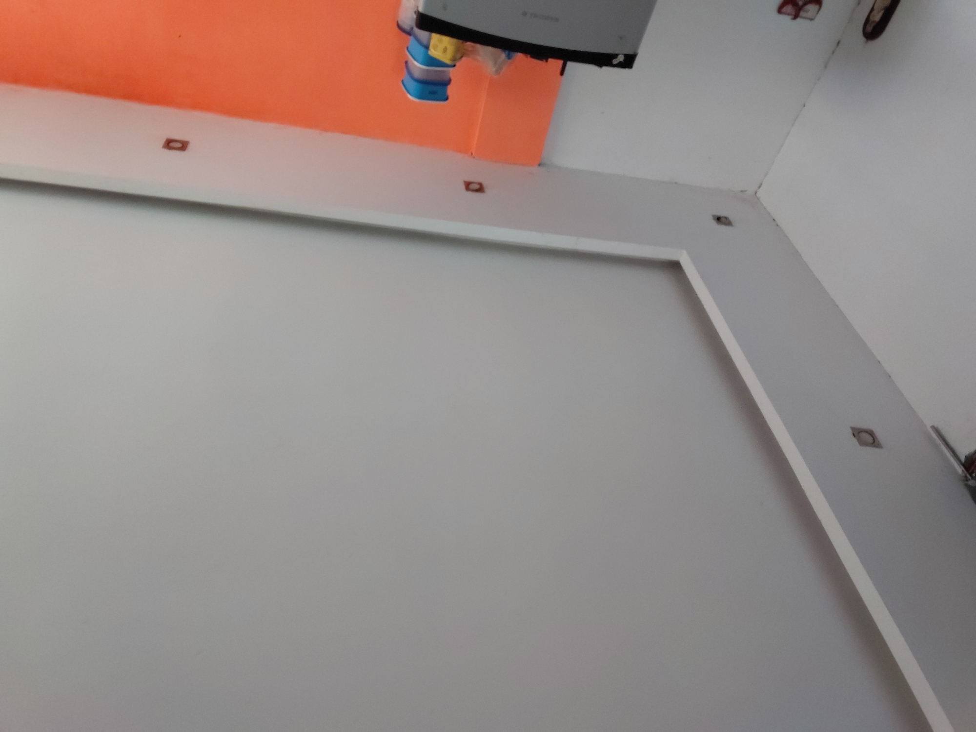 The image captures an interior view of a room, looking upwards from a low angle. The primary subjects are the room's walls and ceiling. A large section of a light grey or off-white wall dominates the lower and left parts of the frame. Above it, an area of a distinct, bright orange wall or ceiling is visible, creating a strong color contrast.
A prominent white beam or architectural molding runs horizontally across the upper part of the grey wall, separating it from the orange section. Several small, square-shaped recessed fixtures, possibly spotlights or sensors, are embedded into this white beam.
Near the top center, suspended from the junction of the orange and white surfaces, is a grey electronic device, possibly an air conditioner unit or a piece of industrial equipment. Beneath this device, some blue and yellow plastic elements or wires are attached. In the upper right corner of the room, where two white wall surfaces meet the ceiling, a reddish-brown electrical box or similar fixture is mounted. Some dark marks and dust are visible along the ceiling lines and corners.
The scene depicts a static indoor environment. There are no people present, and no discernible activity is taking place. The room appears to be functional and possibly contemporary, given the clean lines of the white molding and the contrasting wall colors. The image offers no specific visual cues to determine the time of day or weather, nor does it convey any particular emotions. No clearly readable text is visible on any of the objects or surfaces. The location, Santa Ana, Venezuela, is provided as context but is not visually evident within the image itself.