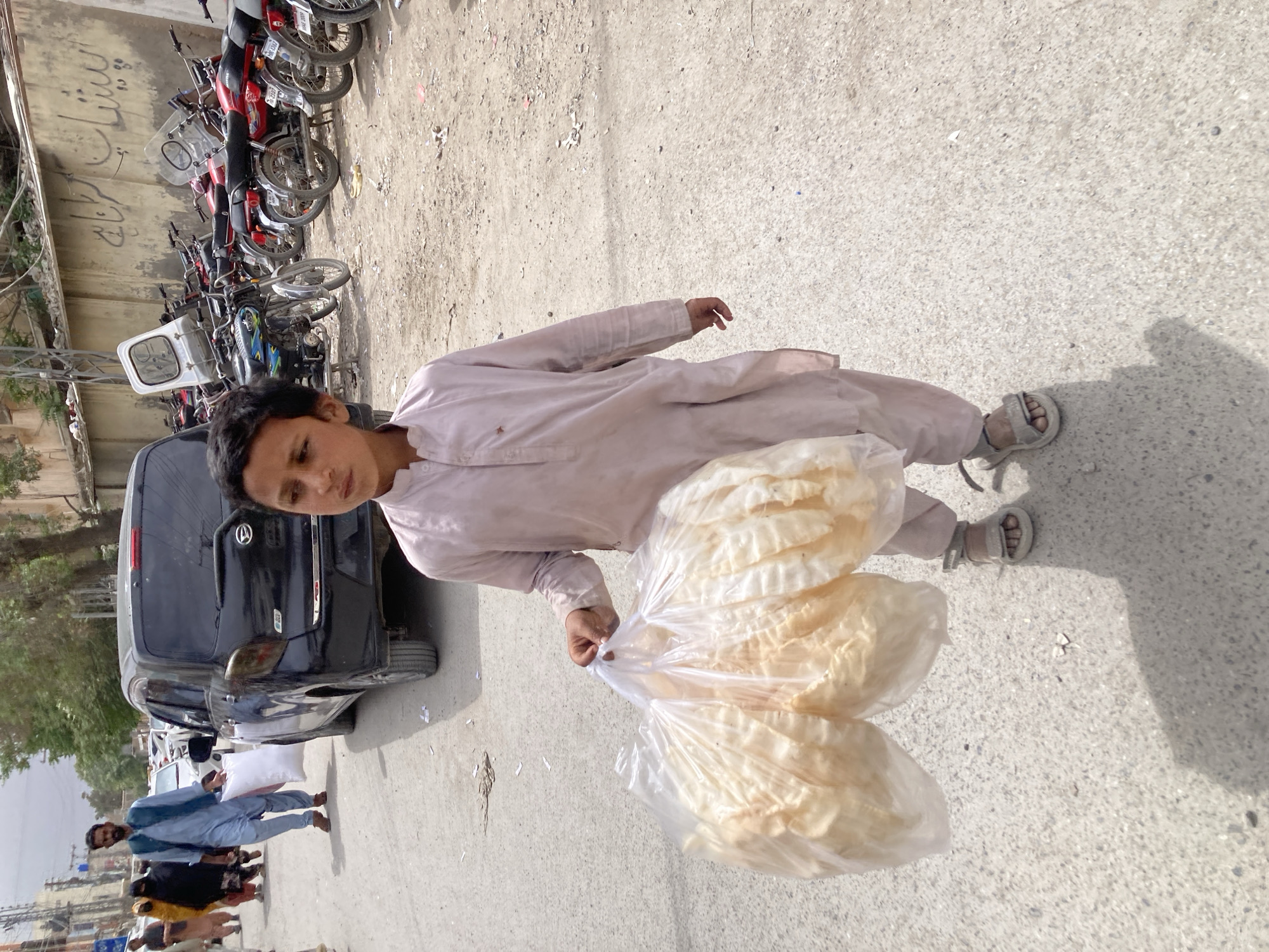 A medium-shot captures a young boy standing on a concrete surface, likely a street or sidewalk. He is dressed in a light-colored, traditional tunic and trousers, and wears sandals. He holds a large bundle of what appears to be puffed rice or wafers, packaged in transparent plastic bags.
In the background, to the left, is a line of parked motorcycles and a black compact car. Further in the background, a small group of people can be seen walking, indicating the presence of a populated area. The scene appears to be in Quetta City Tehsil, Pakistan. The lighting suggests it is daytime.