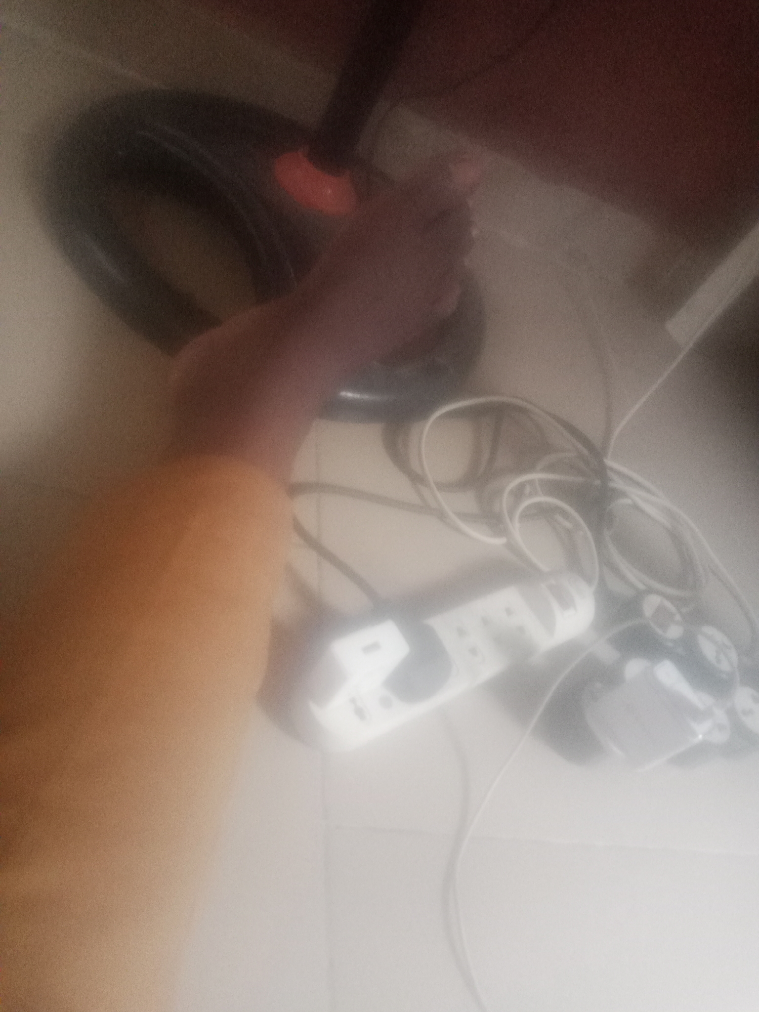 A person's leg, clad in an orange garment, is visible in the lower left of the image. The leg is positioned near a black fan with a red accent on its base. Several electrical cords are tangled around a white power strip, which has multiple outlets. Two gaming controllers, one white and one black with gray accents, are also present. The scene appears to be indoors, possibly in a living area or bedroom, on a tiled floor. The lighting is dim, and the image has a hazy quality.