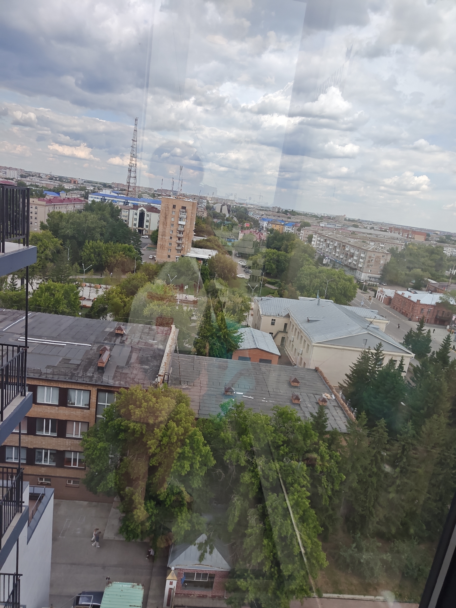 An aerial view captures a cityscape with a mix of residential and industrial buildings under a cloudy sky in Petropavl, Kazakhstan. In the foreground, a modern building with dark railings is partially visible, leading down to a courtyard with two people walking and a green dumpster. Adjacent to this is a brick building with a dark roof and several small structures on top. Further into the city, a variety of multi-story buildings, some with red roofs, are interspersed with lush green trees. A prominent telecommunications tower stands tall in the distance. The reflection of the sky and some unknown objects are visible on a glass surface, partially obscuring the view of the city.