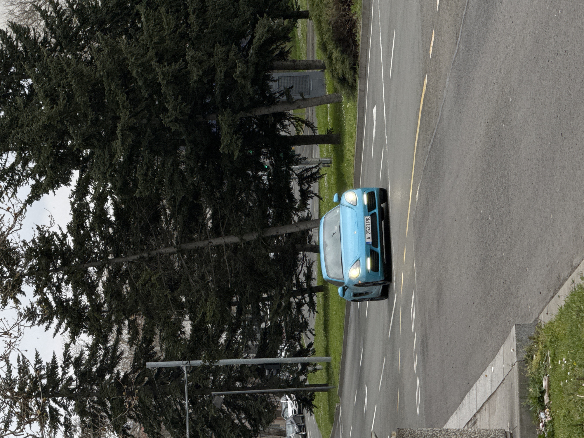 The image shows a light blue car on a paved road, lined with trees and greenery, likely in Burgas, Bulgaria based on the car's license plate ("A" designates Burgas). The road markings are visible, with white and yellow lines. The image is taken from an elevated perspective, and the scene includes urban elements like street lights. No people are visible in the image.