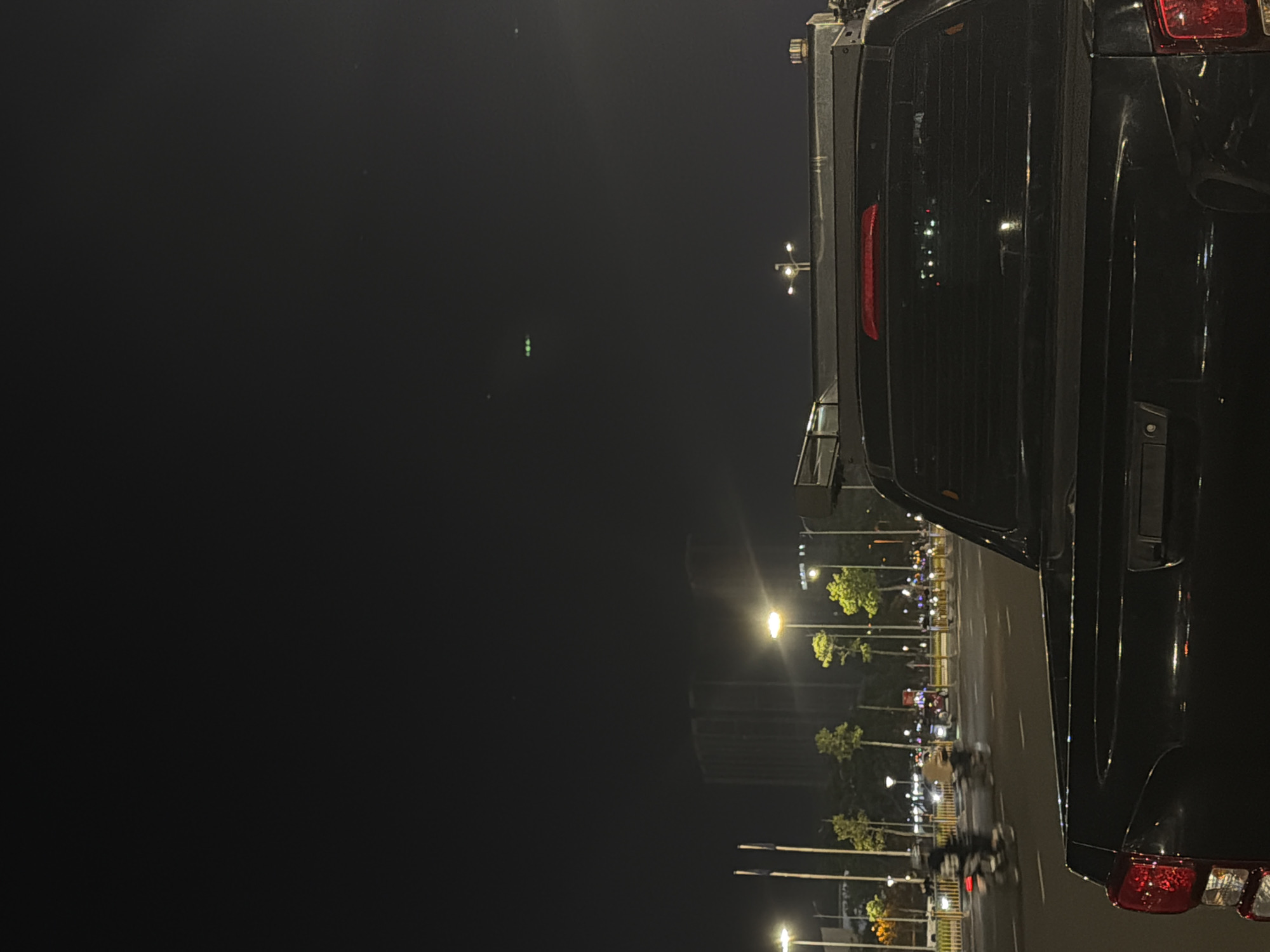 The image shows a nighttime street scene in the city of Đà Nẵng, Vietnam. The foreground is dominated by the rear of a black pickup truck, with its taillights visible on the left. In the middle ground, a street is illuminated by streetlights, with traffic, including several motorcycles, moving along it. Trees line the street, and beyond them, tall buildings, likely skyscrapers, are visible against the dark sky. The sky itself is dark, with a few distant lights that may be aircraft or other aerial phenomena. The overall impression is one of an urban environment at night, with active street life and city infrastructure.