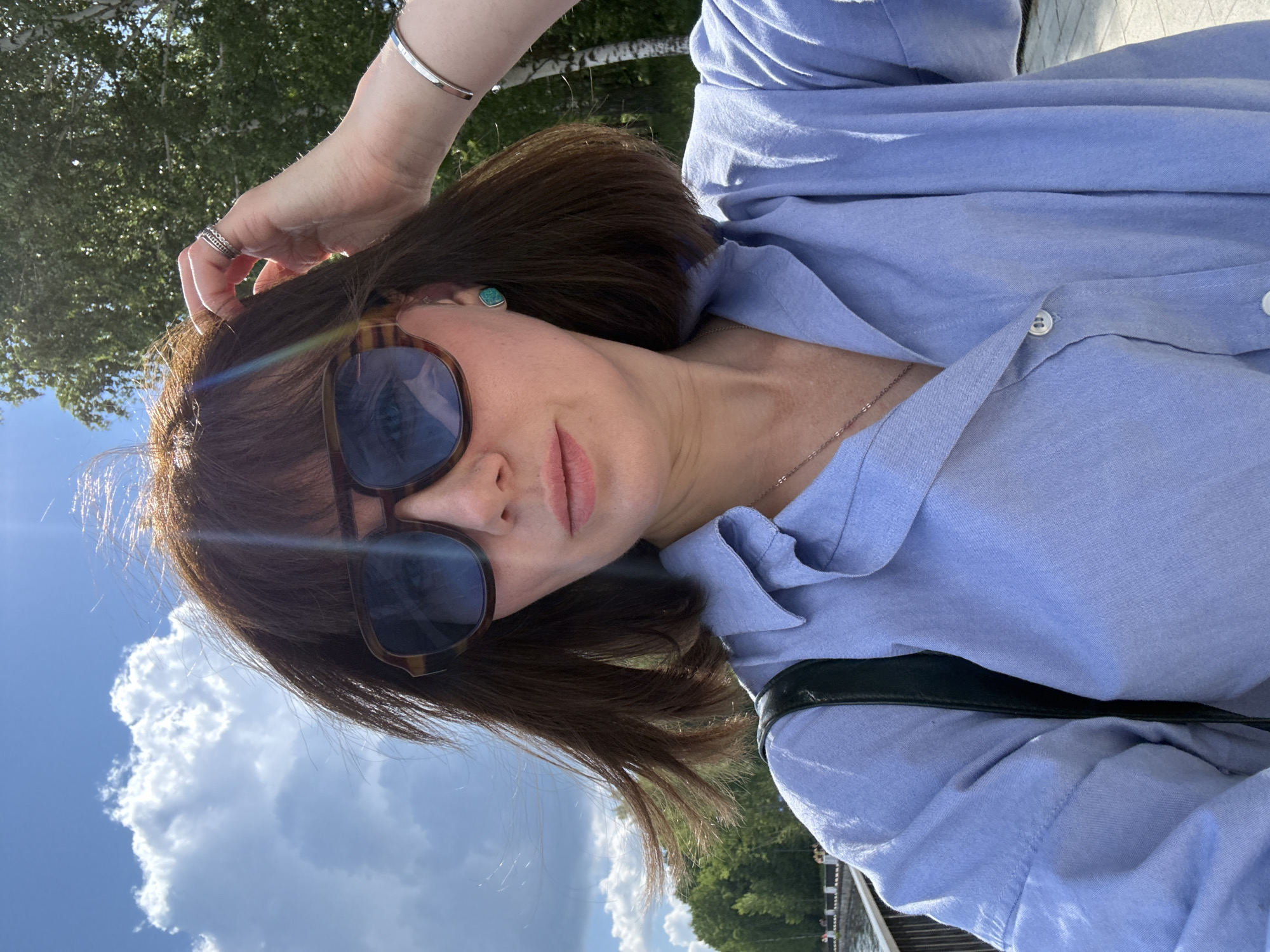 A medium shot captures a woman with shoulder-length brown hair and bangs, wearing large, square-shaped sunglasses with tortoise-patterned frames. She has on a light blue collared shirt, a silver necklace, a silver bracelet, and a silver ring with multiple bands on her right hand. A green bag strap is visible over her left shoulder. She is outdoors with her right hand touching her hair. In the background, there are green trees, a cloudy blue sky, and a body of water with a walkway and railing along the edge, suggesting a location in the city of Chaikovsky, Russia, possibly near the Kama River.