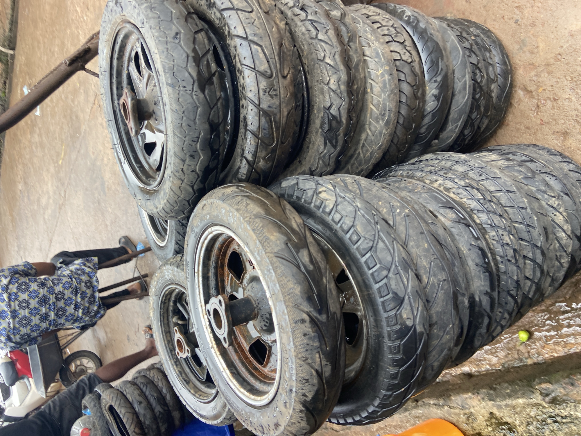 The image displays a collection of used motorcycle tires, some mounted on rims, stacked in an outdoor setting. The tires are black and appear worn, with visible tread patterns. Several individuals are partially visible in the frame, suggesting their involvement in handling or organizing the tires. One person, wearing a blue and white patterned shirt and black pants, is seen from the back, sitting on what appears to be a small cart or vehicle. Another person's arm is visible, reaching towards the tires. The ground is a dirt or concrete surface, wet in places, indicating a possible recent rain or cleaning activity. A small green object, possibly a lime or small fruit, rests on the wet ground near the tires. A metal stand or rack is present in the background, supporting some of the tires. The overall impression is of a tire storage or sale area, likely in a workshop or marketplace. The lighting suggests it is daytime, but there are no strong shadows indicating a specific time. The presence of multiple tires and the apparent activity suggest a commercial or repair-related context. The location is specified as Ogidi, Nigeria, which provides a cultural and geographical context for the scene. There is no discernible text in the image.