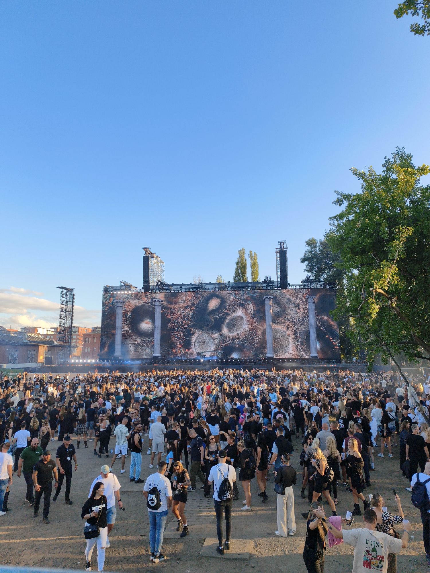 An outdoor concert is taking place in front of a large stage with a screen displaying abstract imagery. A crowd of people, dressed in casual clothing, are gathered on a dirt ground, facing the stage. Several tall towers with lights and equipment are positioned on either side of the stage. The sky above is clear and blue, with a large green tree visible on the right side of the frame. Buildings can be seen in the background, suggesting an urban setting.