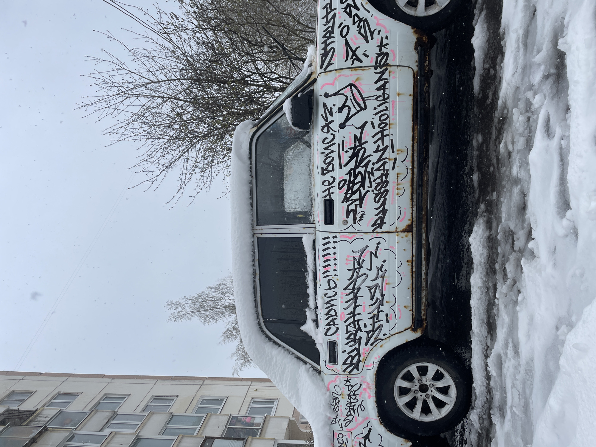 The image captures a winter street scene featuring a white, older model vehicle, possibly a van or mini-bus, parked amidst snow. The car, heavily covered in black and pink graffiti, is seen from its left side, with deep snow accumulated on its roof and along its base, forming a large snowdrift on the adjacent ground. Snow is actively falling, visible as flakes against the uniformly light, overcast sky, contributing to the wintry atmosphere.
The vehicle exhibits signs of age, including rust along its body lines and windows. Its exterior is a canvas for intricate graffiti tags and stylized lettering, predominantly in Cyrillic script. discernible phrases include "НЕ БОИСЯ" (Don't be afraid) near the front window and "САДИСЬ" (Get in/Sit down) towards the rear. The car's wheels feature white multi-spoke rims.
In the background, a large deciduous tree with bare branches occupies the upper left portion of the frame, silhouetted against the sky. To the lower left, a multi-story building with numerous rectangular windows is visible, suggesting an urban or residential setting. The time of day appears to be daytime, though the heavy cloud cover gives a muted light. The scene conveys a sense of cold, quiet, and perhaps neglect, consistent with a snowy day in a city like Moscow, Russia.