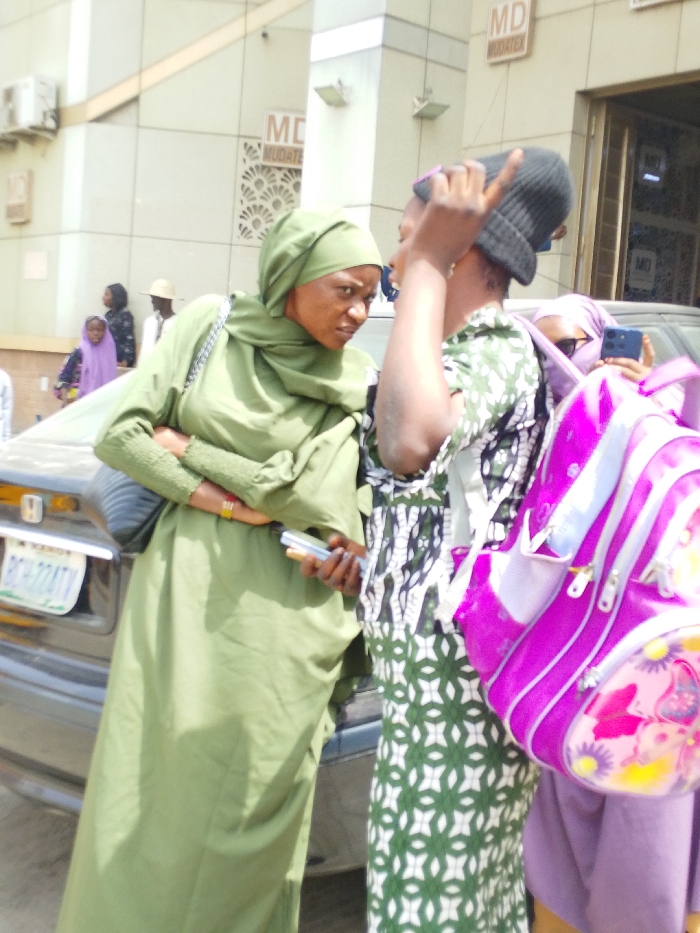 The scene depicts an outdoor urban environment in Sabon Gari East, Kano State, Nigeria, during daylight hours. The primary focus is on three individuals in the foreground. On the left, a woman in a light green hijab and matching long-sleeved dress holds a white smartphone in her right hand, her left arm crossed over her midsection; she faces towards the right with a serious expression. Adjacent to her, a second woman, partially visible from the chest up, wears a black knit beanie and a white and dark green patterned dress, with her right hand raised and fingers curled as if in mid-gesture or speech. Behind this second woman, a third individual, partially obscured, wears a light purple hijab and holds a dark-colored smartphone, directed towards the scene. A bright pink backpack with multi-colored floral designs is prominently displayed in the lower right foreground. Behind the woman in green, a dark-colored sedan is partially visible, displaying a Nigerian license plate: "KANO PCH224TV." The background features a light-colored building with windows and air conditioning units, bearing multiple "MD MUDATEX" signs. Further in the distant left background, additional individuals, including children and a man wearing a light hat, are visible.
