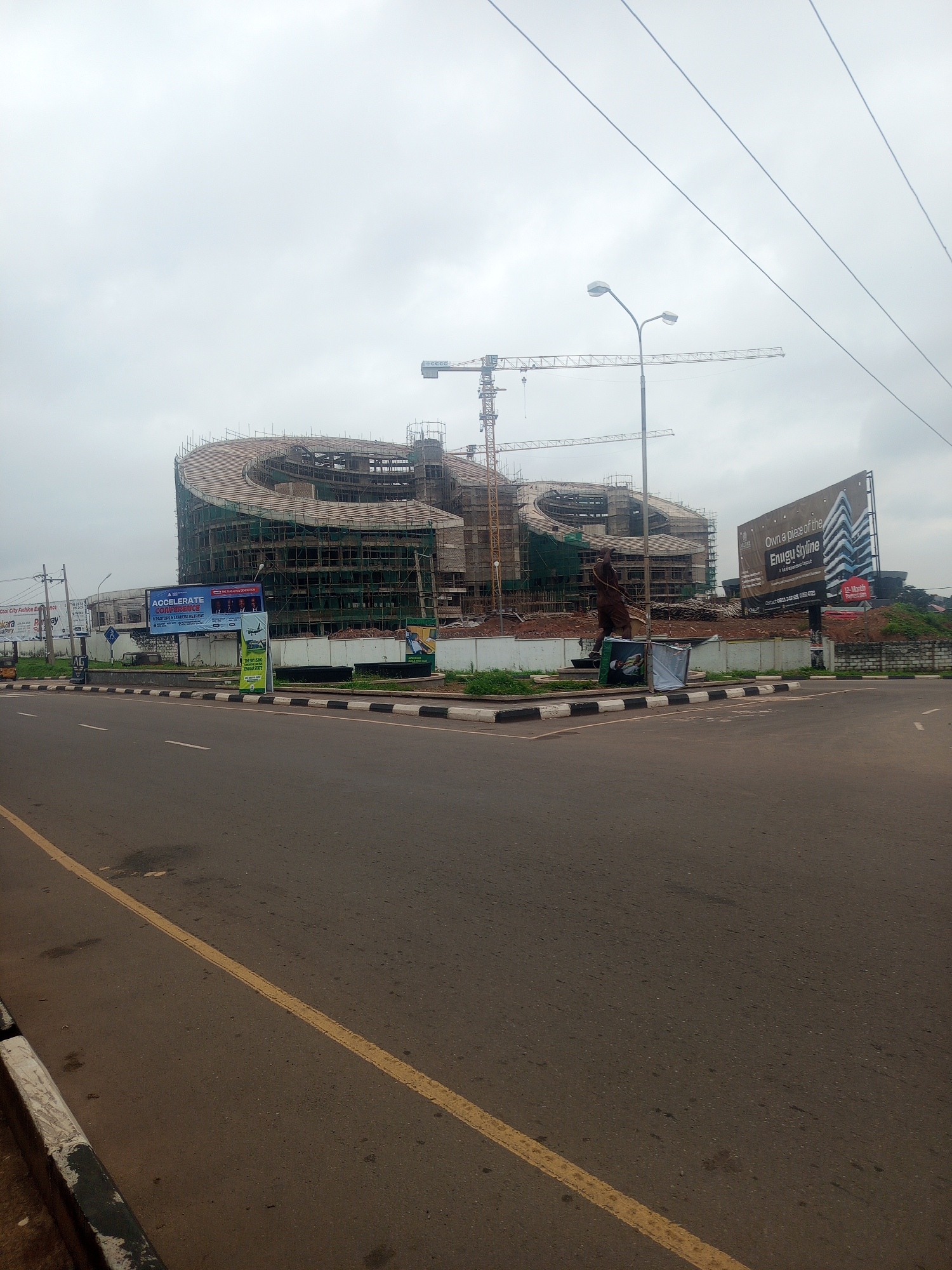 This image depicts a large, unfinished building under construction in Enugu, Nigeria. The building has a distinctive curved, spiral design and is surrounded by scaffolding. Two large cranes are visible, indicating ongoing construction work. In the foreground, a road with yellow lane markings runs past the construction site.
Several billboards are positioned along the road. One prominent billboard advertises an "ACCELERATE CONFERENCE" with the subtitle "PASTORS & LEADERS RETREAT." Another billboard features an artist's rendition of a modern building complex with the text "Own a piece of the Enugu Skyline." A smaller green billboard states "THE SKY'S NO... ENUGU STATE." A statue of a person is also visible near the construction site.
The weather appears overcast, suggesting a cloudy or possibly rainy day. The time of day is not explicitly indicated, but the general lighting suggests it could be daytime. The scene is urban, with the road and billboards indicating a public space in a city. The overall impression is one of development and progress, with a focus on a major construction project. The image captures a moment in the transformation of the urban landscape of Enugu.
