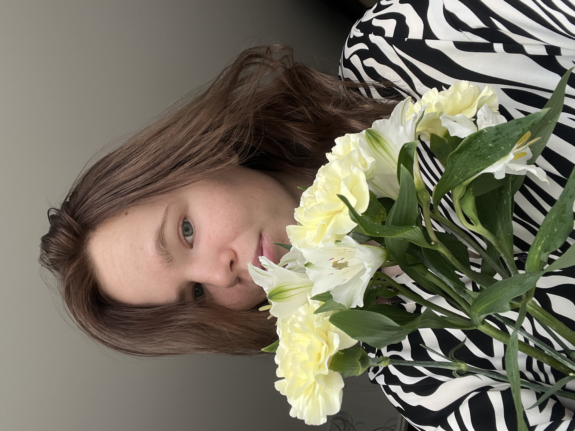 An image, presented rotated 90 degrees clockwise, shows a young woman in Chaikovsky, Russia, holding a bouquet of flowers. Her head is on the left side of the frame, and she looks towards the viewer with a neutral expression. She has light brown hair, which falls around her face and shoulders, and light-colored eyes.
She is holding a bouquet consisting of pale yellow carnations and white alstroemerias, characterized by their green stems and leaves. The flowers are prominently displayed in the center and right side of the image. The woman is wearing a garment with a distinct black and white zebra print, visible on her right side, partially covered by the flowers.
The background is a plain, light grey wall, indicating an indoor setting with soft, even lighting. There is no visible text in the image.