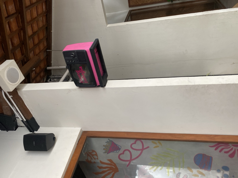 The image shows the exterior of a building in Surakarta, Indonesia, located at the coordinates -7.557 latitude and 110.796 longitude. A pink, faux vintage television, mounted to a white wall, with the words "Ruang Bahagia" (which means "Happy Room") written on it, is the focal point. The TV is mounted to a wall, which is part of a corner of the building. Another wall meets the corner, and an open doorway with a wooden frame leads into a space with a white wall. To the left of the television, a wooden door frame, leading into another space, has a glass panel that displays colorful artwork on it. In the background, a ceiling with a tiled, brown surface is visible above the doorway and the television. A white ceiling speaker can be seen in the upper left of the image. There are no people present in the image.