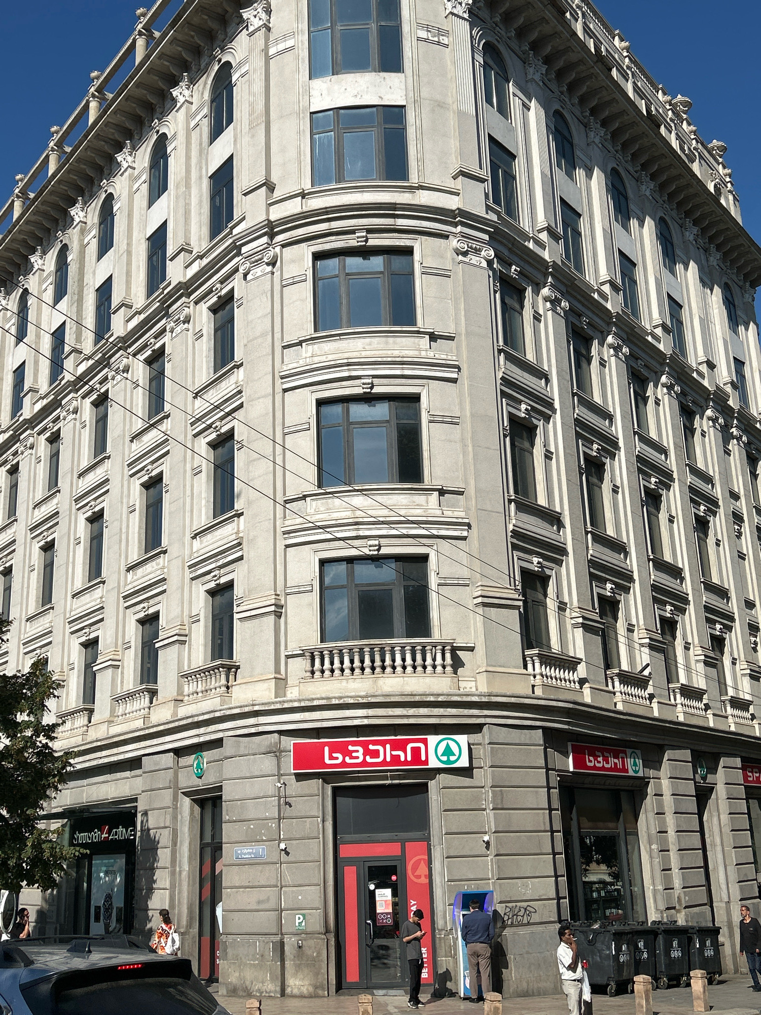 The image depicts a large, ornate, light-colored stone building with multiple stories and arched windows. The building has a classic architectural style with columns and decorative elements along the roofline and around the windows. On the ground floor, there are several storefronts, including one with a red sign that reads "SPAR" in Georgian script and a green circular logo. Another storefront displays a sign with "showtime" and "4+time" in English. Several people are visible on the sidewalk in front of the stores. A blue ATM is also present. To the left of the building, a tree with green foliage is partially visible, and a dark-colored car is parked on the street in the foreground. The sky is clear and blue, indicating a sunny day. The presence of the SPAR store and the Georgian script on the signage strongly suggests the location is in Tbilisi, Georgia.