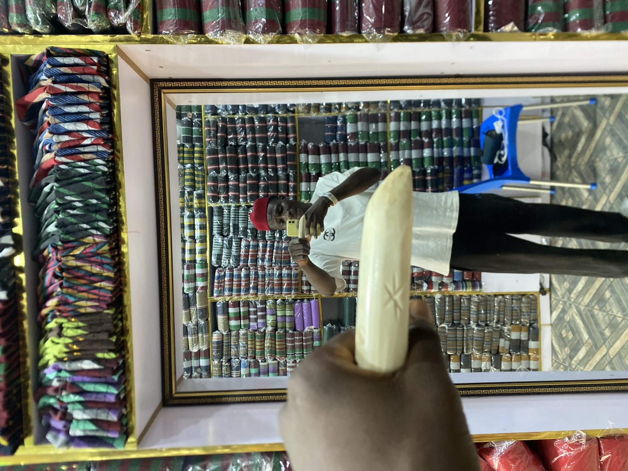 The photograph shows a person holding an object, possibly a tusk, in front of a mirror within a shop in Bukuru, Nigeria. The reflection shows a man in a white shirt and red cap taking a photo of himself with a yellow cell phone. The shop displays stacked fabrics of various colors and patterns. Above the mirror are bundles of fabric wrapped in plastic. A blue chair is visible in the reflection.