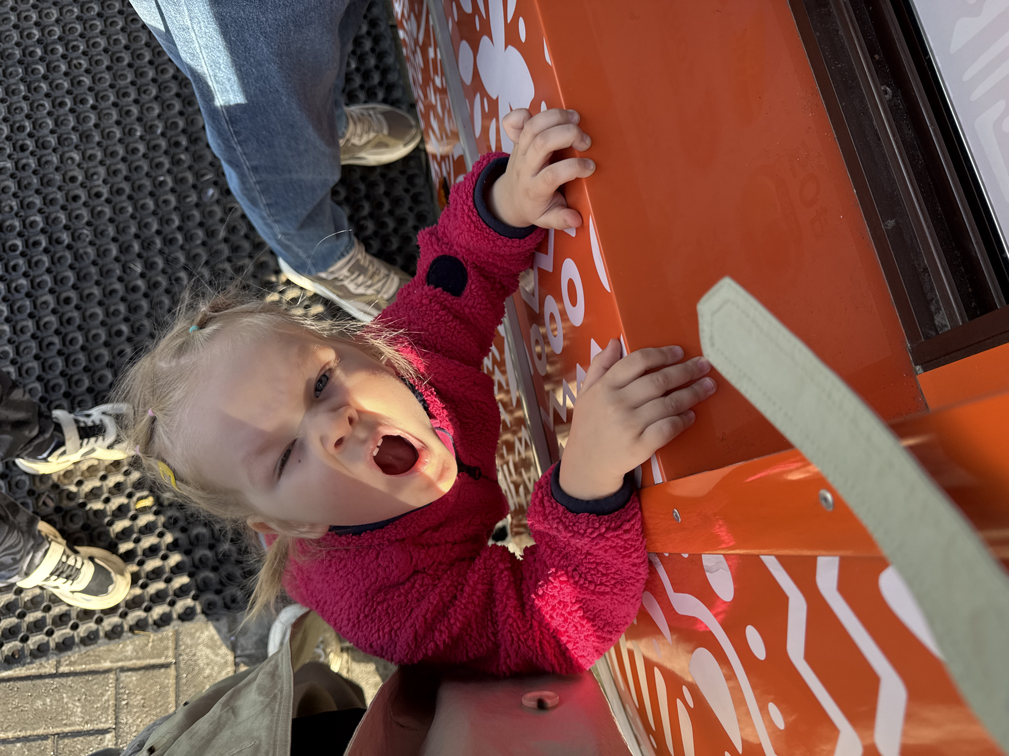 A child with blonde hair, wearing a pink jacket, reaches up to touch a decorated orange surface while partially opening their mouth and winking. The scene is framed from an elevated, slightly canted perspective, capturing additional adults nearby. The location is identified as Perm, Russia. The surface is embellished with white patterns. The child grips the orange surface with both hands. The jacket is fleece. One adult is wearing jeans and tennis shoes and another is wearing tennis shoes and an olive-green jacket. A black and patterned mat can be seen under the feet of the adults and child.