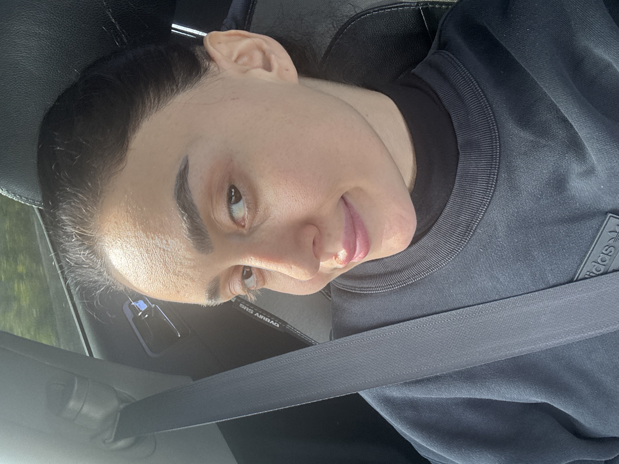 A light-skinned person with dark hair is seated in a car, wearing a black Adidas shirt and a seatbelt. The car's interior is visible, including part of the dashboard and the SRS AIRBAG tag on the seatbelt. The location is purportedly in the city of Tbilisi, Georgia, but there is no visual confirmation of this in the image. The person has a small wound or irritation on their lower lip.