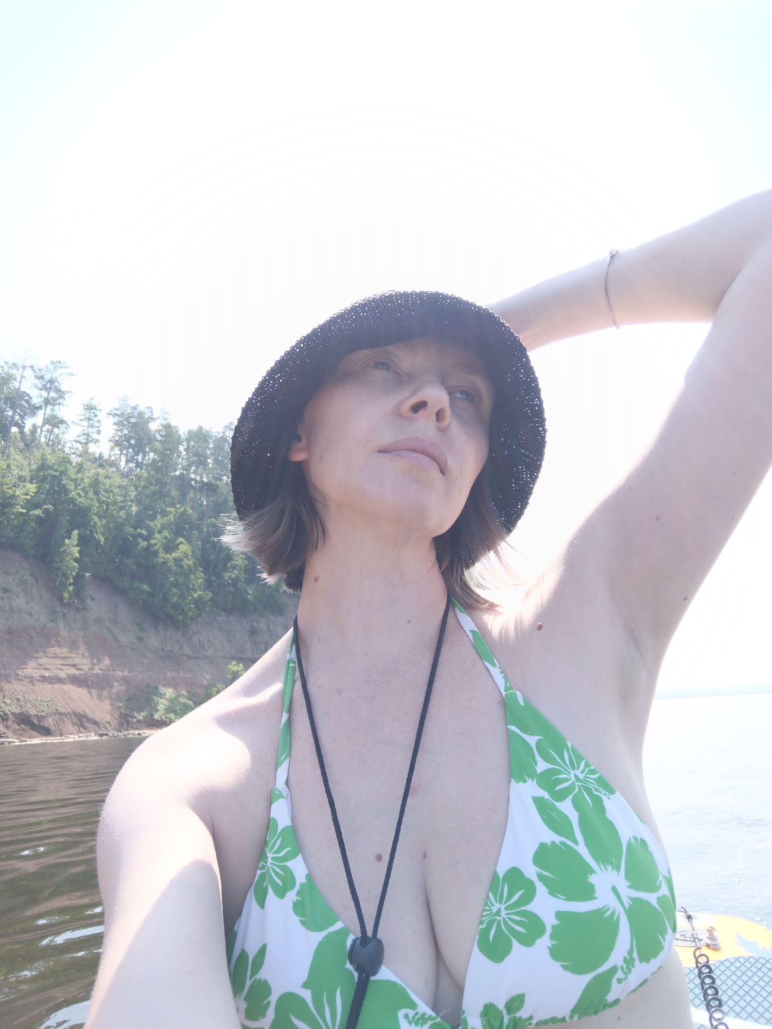 In Noviy, Russia, a woman is captured in a selfie on the water, possibly on a boat or paddleboard, with a steep, tree-covered bank in the background. The sun is high, creating a bright, hazy sky. The woman wears a black bucket hat and a green and white floral bikini. Her right arm is raised, hand resting on her head, and a delicate bracelet adorns her wrist. The water is calm and reflects the light.