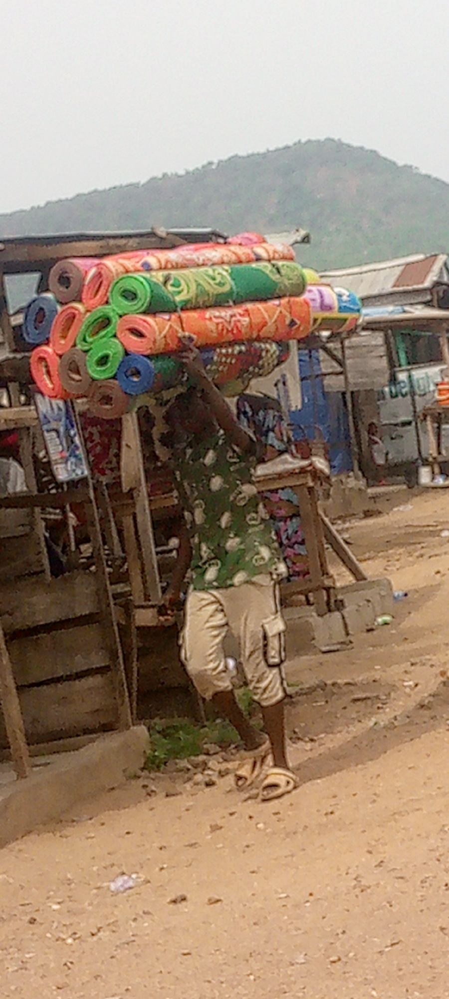 The image depicts an outdoor street or market scene in Adankolo, Nigeria. The primary subject is a man walking on a dirt path, carrying a large, towering stack of rolled mats on his head. The mats are brightly colored and patterned, appearing to be prayer mats or sleeping mats, meticulously arranged and secured for transport.
The man is dressed in a green short-sleeved shirt with a light pattern, light-colored cargo shorts, and light-colored sandals. His left arm is raised, suggesting he is steadying the substantial load on his head.
The background reveals simple wooden structures, likely market stalls or shelters, indicating a commercial environment. A prominent green-covered hill or mountain rises in the distance under a hazy, overcast sky, suggesting daytime with subdued lighting. To the right, a white vehicle, possibly a small delivery truck or tricycle, is partially visible, with the word "deligh" or "delight" faintly readable on its side. The ground consists of reddish-brown dirt, characteristic of unpaved roads in many parts of the region. The scene captures an everyday activity of a vendor transporting goods in a bustling, semi-urban or rural setting.