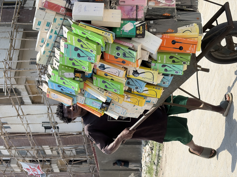 In Igboefon, Nigeria, an adult male street vendor pushes a single-wheel metal wheelbarrow laden with electronics merchandise forward on an unpaved road under bright sunlight.
The vendor, with dark skin and short, dark, unkempt hair, wears a dark t-shirt, dark green shorts, and olive-green slide sandals. He grips the wheelbarrow's handles with both hands, his gaze directed forward. The wheelbarrow features an attached metal display rack showcasing a dense arrangement of products. Merchandise includes numerous packaged wired earphones (in colorful boxes, primarily green, blue, yellow, and orange, with brands such as "DEEPER BASS," "JINDARX," "PROMAC," and "FRESH"), various white power adapters/chargers (some labeled "FAST CHARGER" or "9A"), and an assortment of plastic phone cases (clear, dark, and some pink).
In the mid-ground to the left, a second male figure stands near a light-colored concrete wall, appearing to look down at a mobile device. The background features a multi-story building under construction or renovation, enveloped by extensive bamboo scaffolding. A red banner with partially visible white text ("ONG") hangs from the scaffolding. The ground is a light-colored dirt or gravel surface, marked by shadows from the wheelbarrow and vendor.