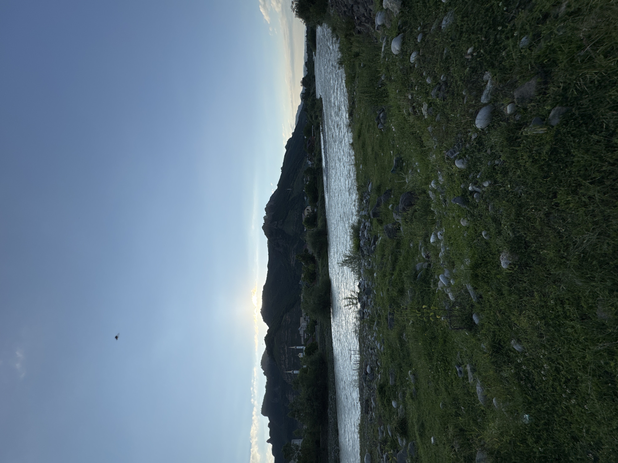The image depicts a serene natural landscape with a wide river flowing through the foreground. The river's surface is rippled, indicating a gentle current. On the riverbanks, lush green grass and scattered rocks create a natural, untamed setting. In the background, a range of dark, mountainous terrain rises, with the setting sun casting a warm glow and silhouetting the peaks. A few buildings, including structures with minarets that suggest a mosque or religious complex, are visible nestled within the foothills. The sky above is a clear, pale blue, with a few wispy clouds, and a small, dark object, possibly a drone or bird, is visible in the upper left quadrant. The overall atmosphere is tranquil and picturesque, capturing the natural beauty of the region. This scene is located in Karachayevsk, Russia, a city known for its mountainous setting and proximity to the Caucasus Mountains.