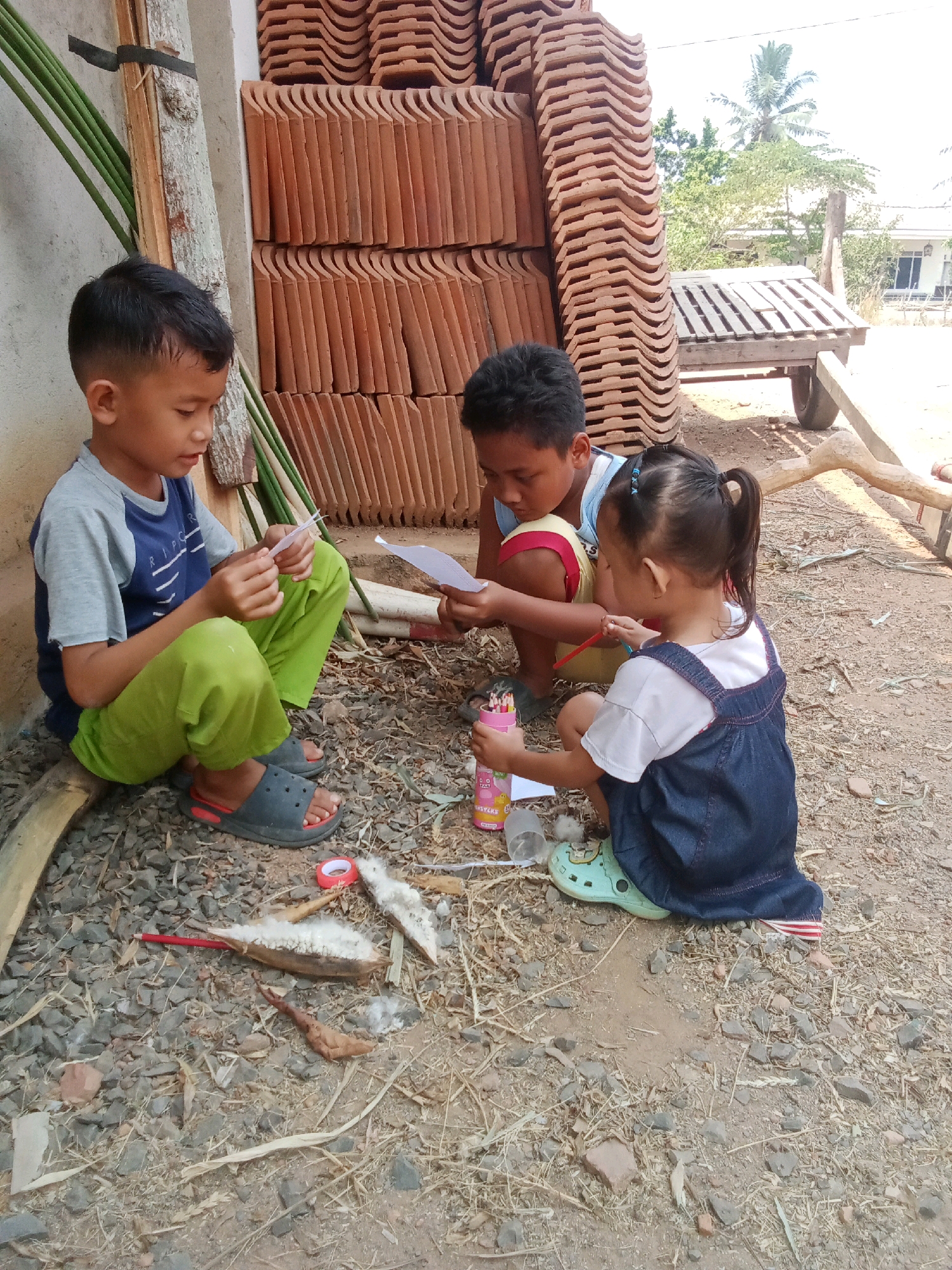 Three children are sitting on the ground outdoors, engaged in an activity. The child on the left, a boy wearing a blue and grey t-shirt and green pants, is holding a piece of paper. The child in the middle, another boy, is also holding paper and appears to be interacting with the child on the right. The child on the right, a girl with her hair in a ponytail and wearing a denim dress over a white t-shirt, is holding a pencil and a container of colored pencils. The ground is covered in dirt, gravel, and dry vegetation. In the background, there are stacks of terracotta roof tiles, some bamboo poles, and a wooden cart. The setting appears to be a rural or semi-rural area, possibly a construction site or a backyard.