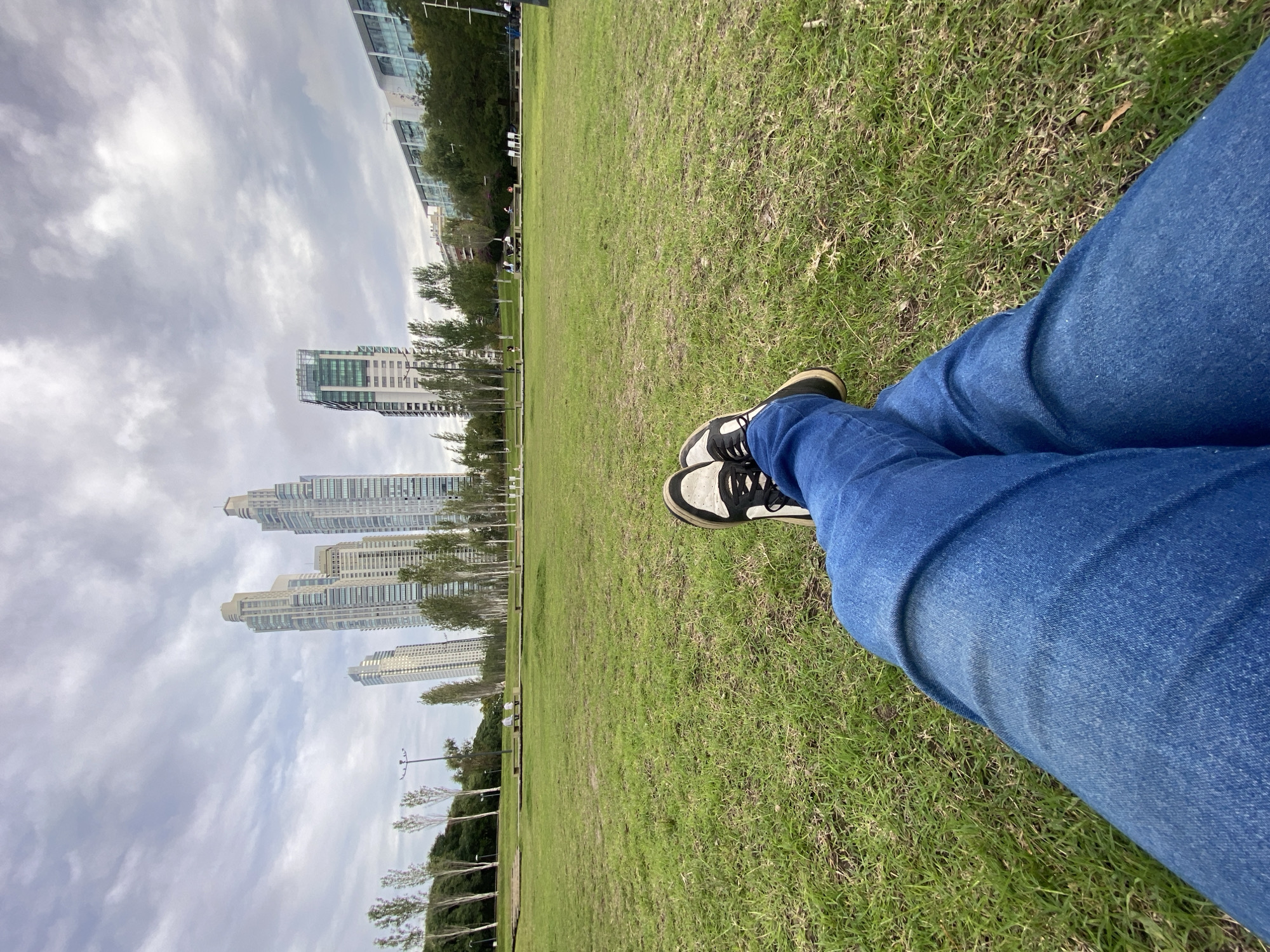 A person wearing blue jeans and black and white sneakers is sitting on a grassy lawn. In the background, there is a view of modern buildings and skyscrapers, a characteristic feature of Puerto Madero in Buenos Aires, Argentina. The sky is overcast with clouds. The grass is green and slightly overgrown.