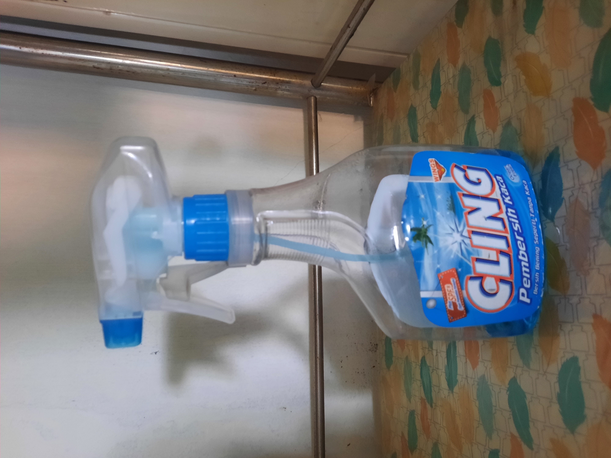 A partially filled, clear plastic spray bottle with a blue trigger nozzle and blue cap stands on a patterned surface. The bottle contains a light blue liquid. The label on the bottle is predominantly blue with white text that reads "CLING" in large letters. Below this, smaller text reads "Pembersih Kaca" and "Bersih Bening Seperti Tanpa Kaca". A smaller red label with the word "SPECIAL" is visible near the top of the main label. The background is a textured, light-colored wall. The bottle is positioned in the foreground, occupying the center of the image, and appears to be an indoor setting. There is no information in the image to suggest it is located in the city of Lhokseumawe, Indonesia.