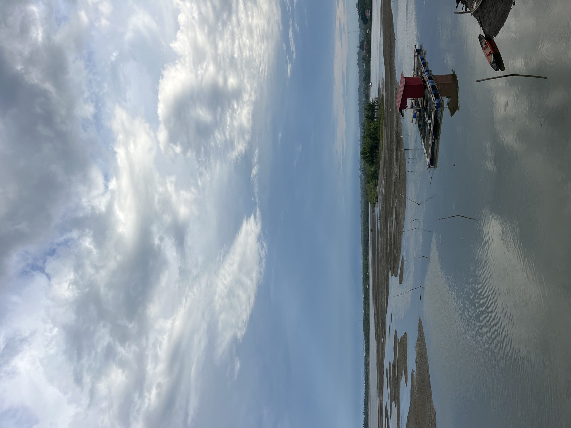 The image depicts a waterscape under a partly cloudy sky. A structure with a red roof sits on the water, possibly a floating platform or fish farm. A small boat is nearby. The water reflects the sky and surrounding area. The shoreline is visible, appearing as a mudflat with scattered vegetation. While I cannot confirm the location, Lhokseumawe, Indonesia is mentioned. If in Lhokseumawe, the image suggests a coastal or riverside scene with human activity related to water, such as fishing or aquaculture. The image lacks people, focusing on the built and natural environment.